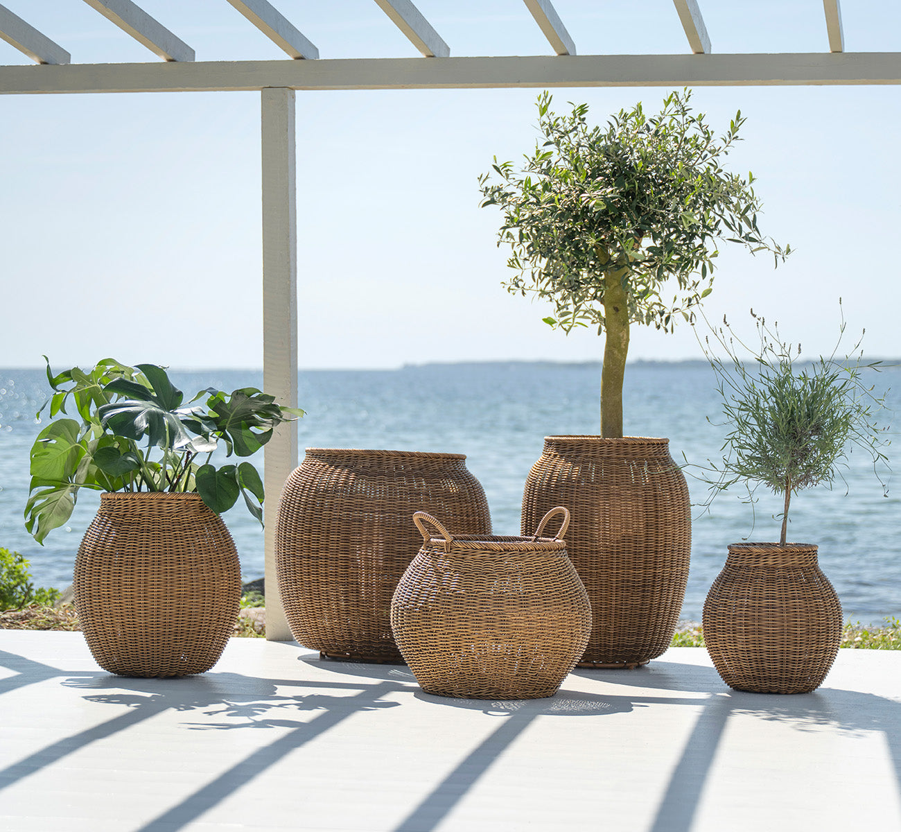 This is a lifestyle image of Alma Basket in chestnut core weave ArtFibre Aluminium, outdoors on a sun-lit patio with a white wall and tile flooring.