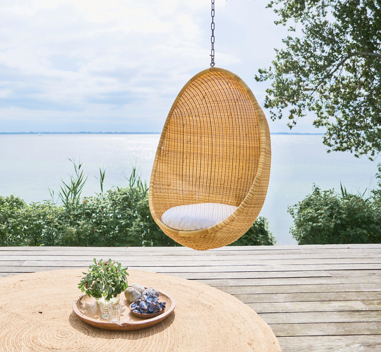 This is a lifestyle image of Hanging Egg Exterior Chair in natural core weave ArtFibre Aluminium, hanging from a white pergola with sandy beach and.