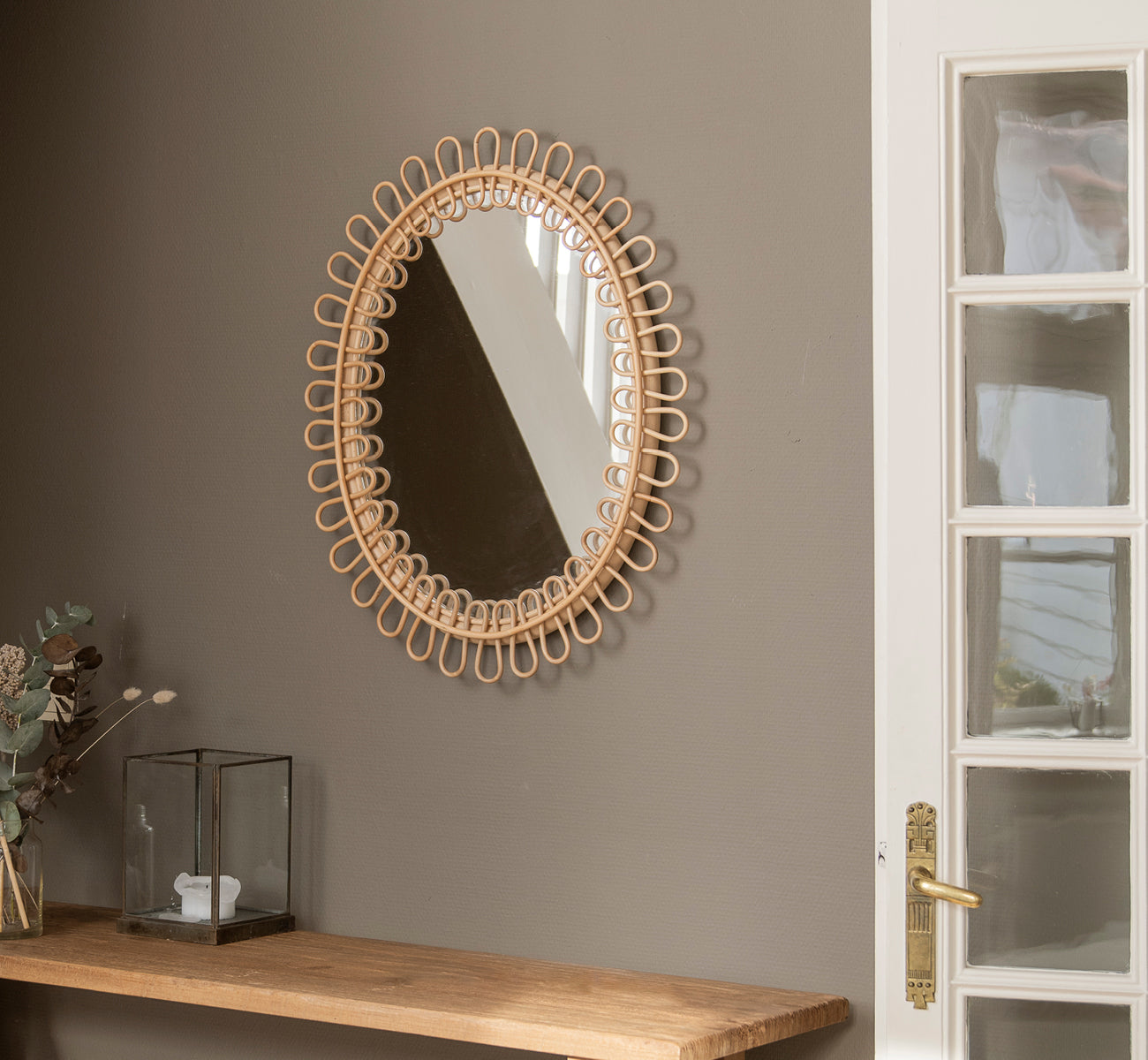 This is a lifestyle image of Luella Mirror in polished natural rattan. Hanging on a gray wall above a wooden shelf with eucalyptus and glass décor.