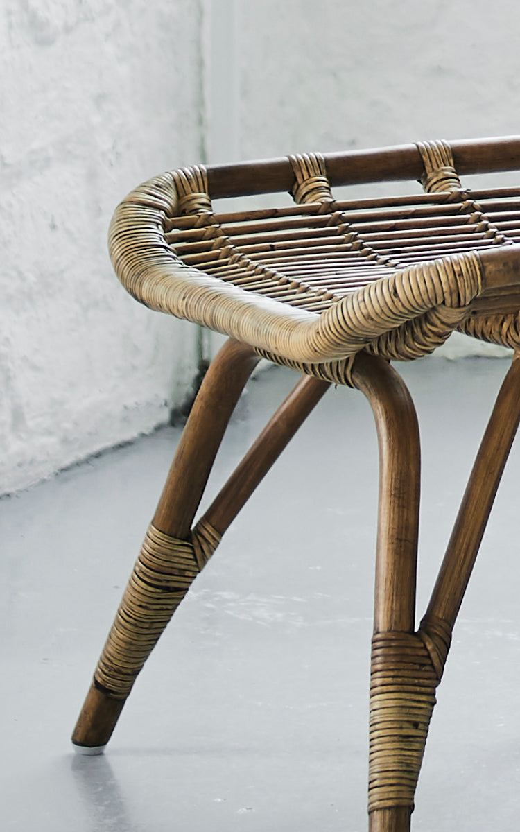 This is a close-up image of Monet Footstool in antique rattan. Detail of the flat woven seat surface and splayed rattan legs.