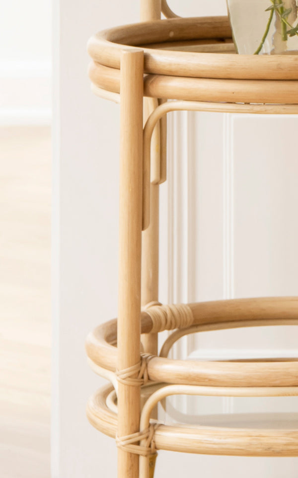This is a close-up image of Romeo Trolley in natural rattan. Detail of the two-tier rattan frame with glass shelf and brass wheels.