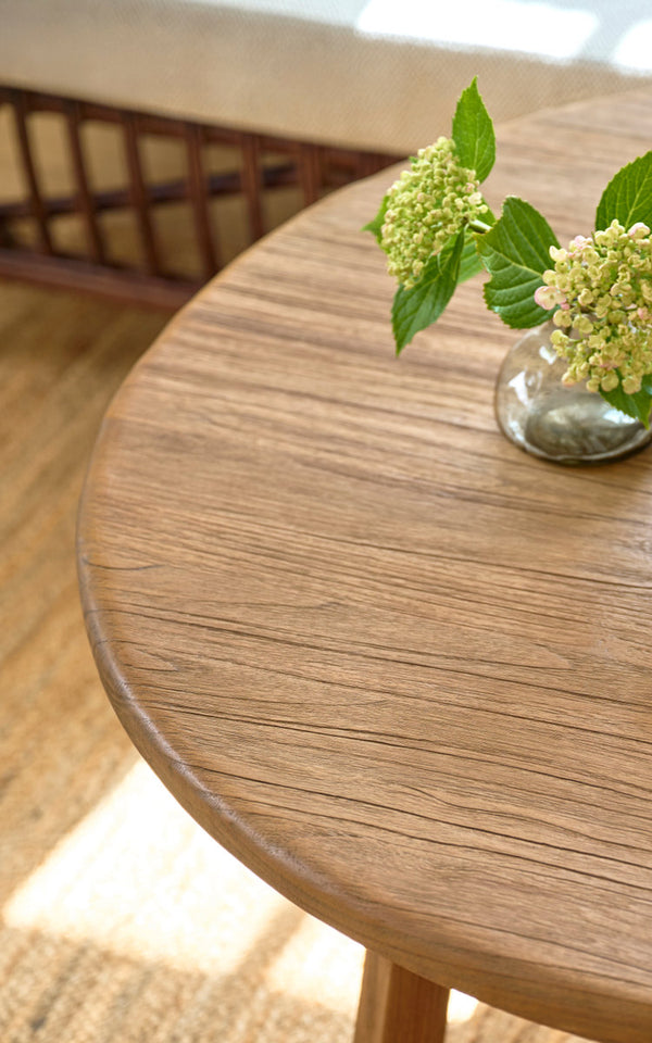 This is a close-up image of Aksel Coffee Table Ø100 cm in teak indoor. Round teak top with hydrangea vase on a jute rug.