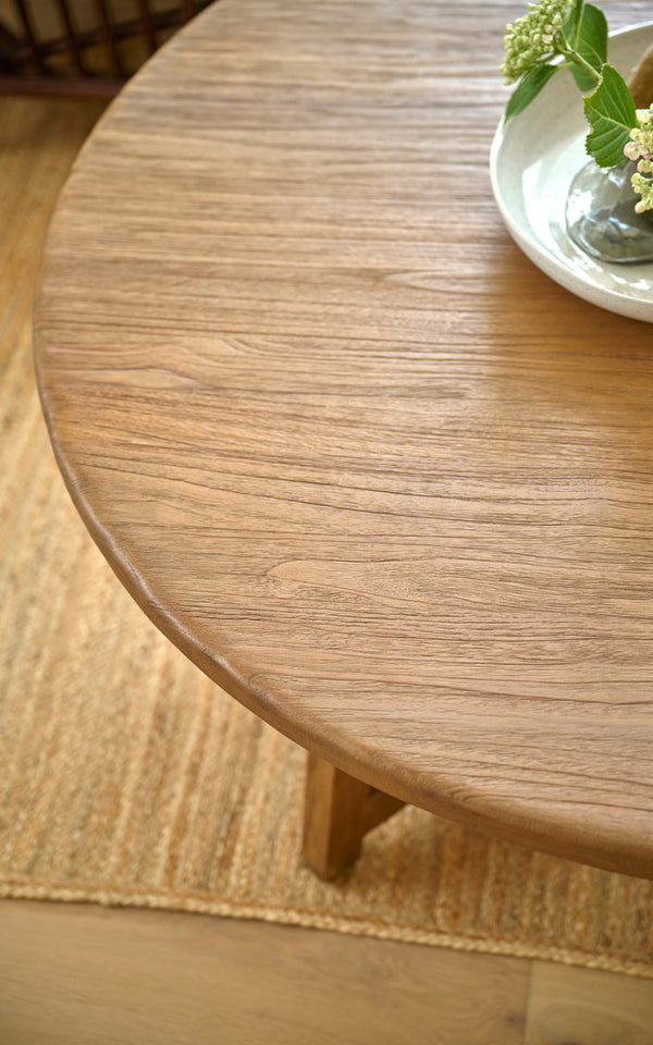 This is a close-up image of Aksel Coffee Table Ø120 cm in teak indoor. Round teak top with flowers in a bowl on a jute rug.