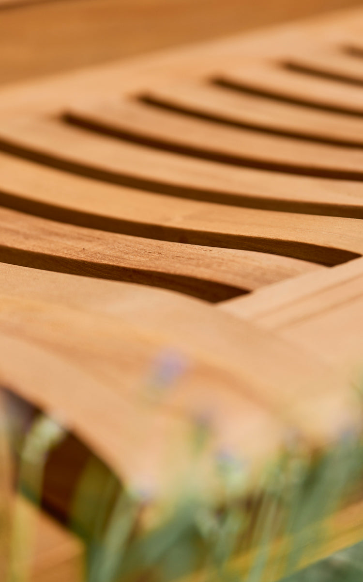 This is a close-up image of St Catherine Teak Bench in teak outdoor. Detail of the curved teak slats of the bench seat surface.
