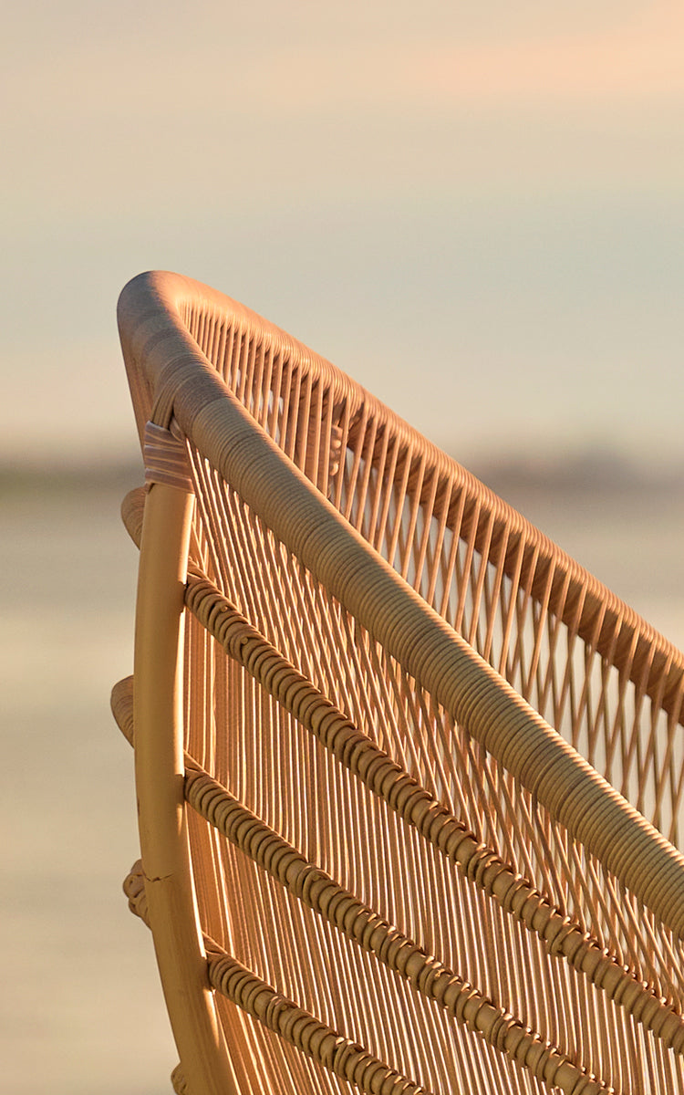This is a close-up image of Luna Exterior Lounge Chair in natural aluminium artfibre. The woven backrest glows in golden sunset light by the sea.
