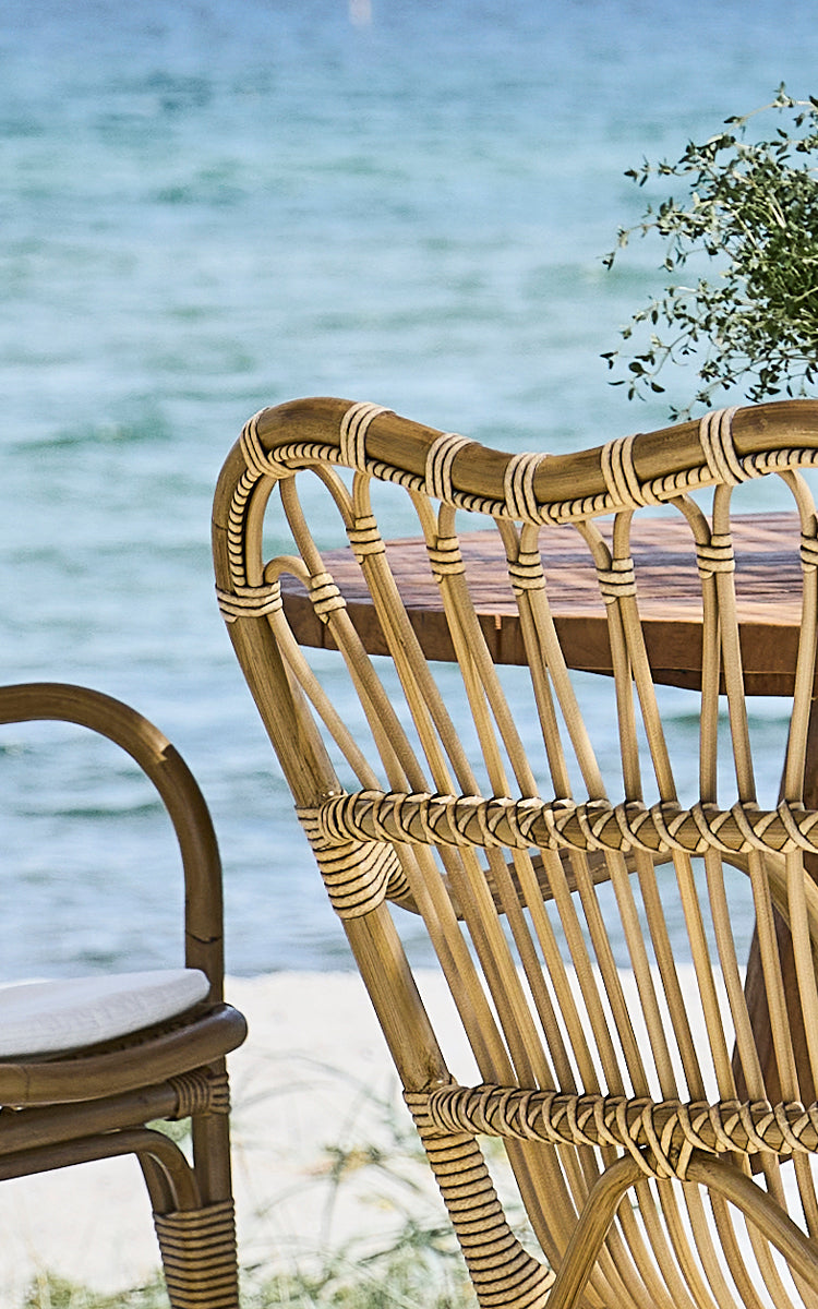 This is a close-up image of Margret Exterior Dining Armchair in antique aluminium artfibre. Curved backrest with wrapped ArtFibre and ocean backdrop.