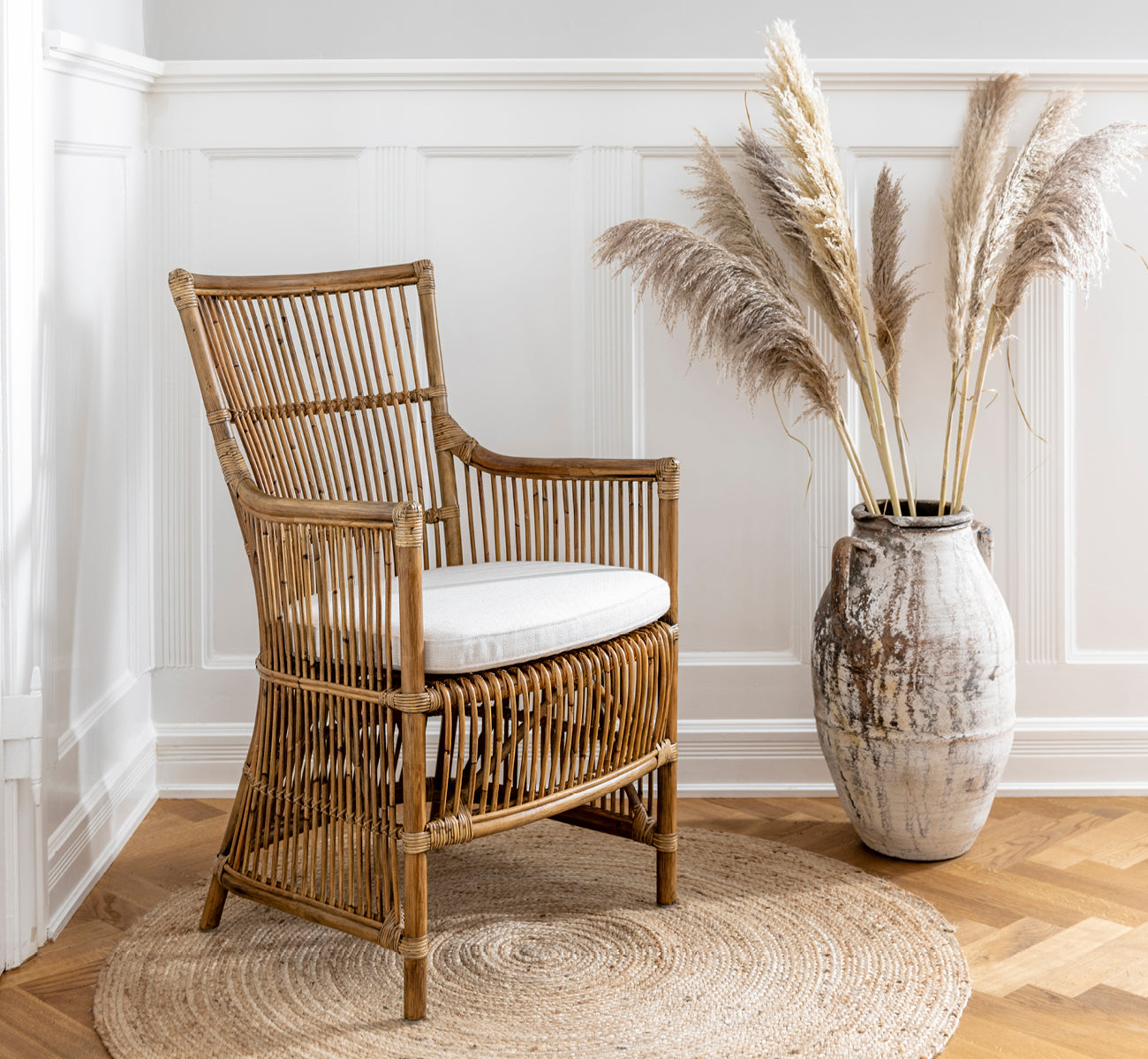 This is a lifestyle image of Davinci Dining Chair in antique rattan, in a bright interior with white panelled walls and herringbone parquet.