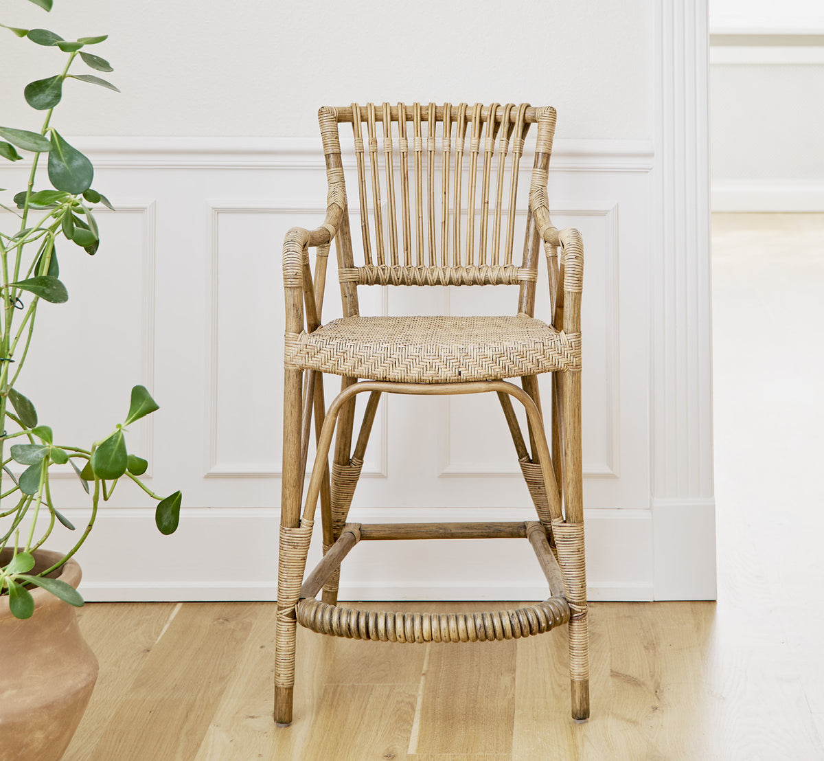 This is a lifestyle image of Blues Counter Stool in antique Rattan, in a bright interior with white panelled walls and herringbone parquet.