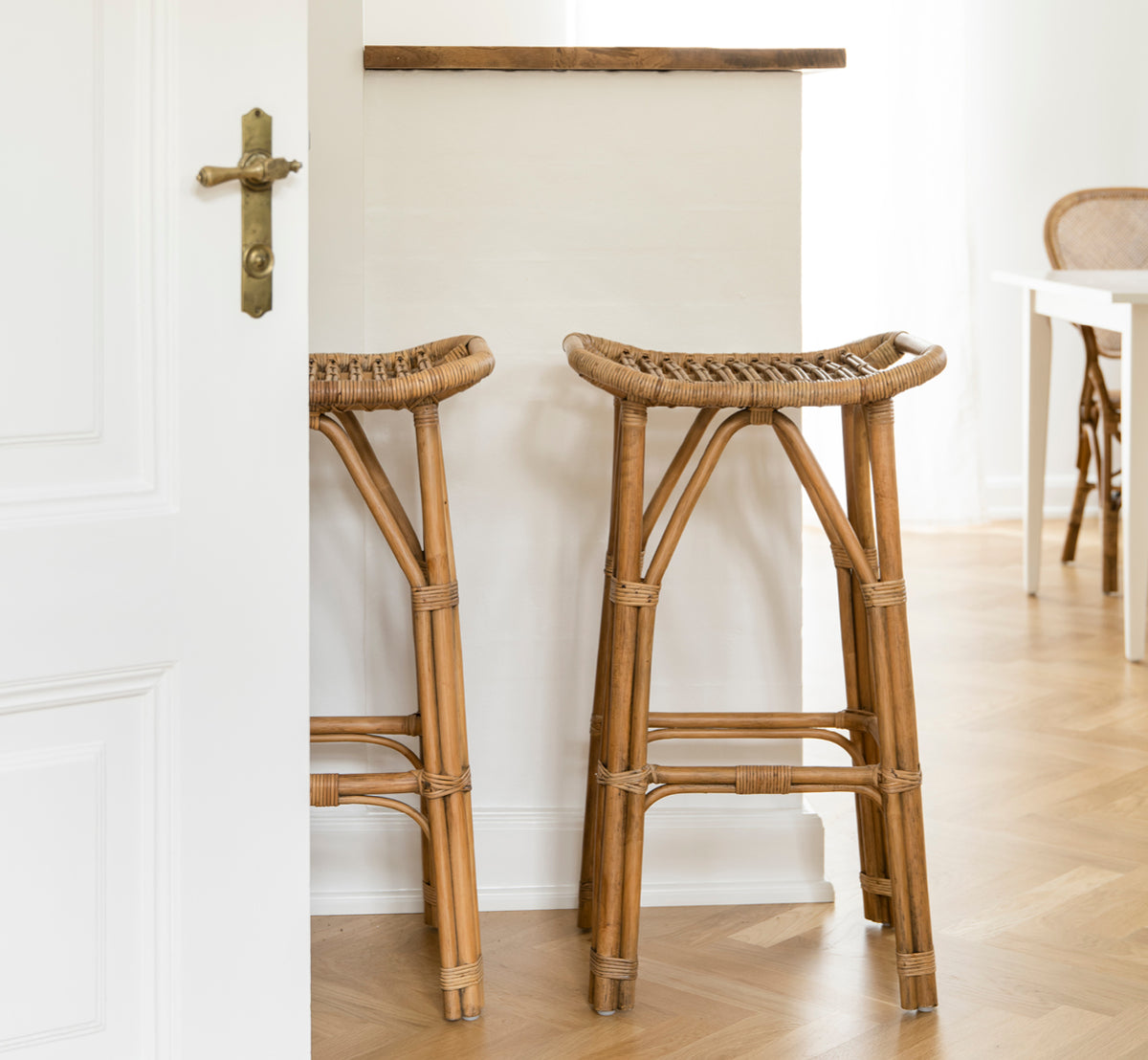This is a lifestyle image of Salsa Bar Stool in antique rattan. A pair placed against a white wall on a herringbone parquet floor.