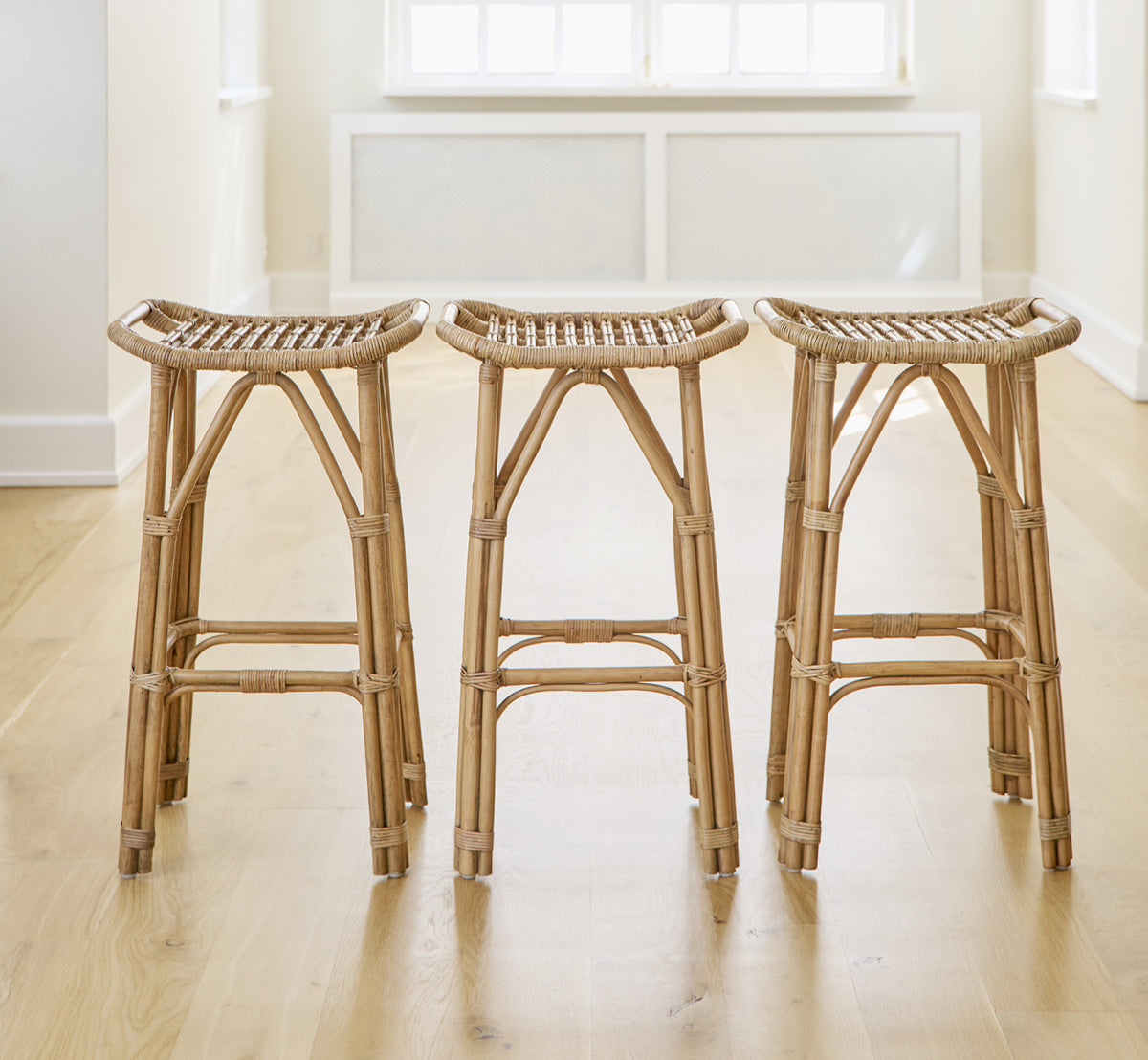 This is a lifestyle image of Salsa Bar Stool in antique rattan. A pair placed against a white wall on a herringbone parquet floor.