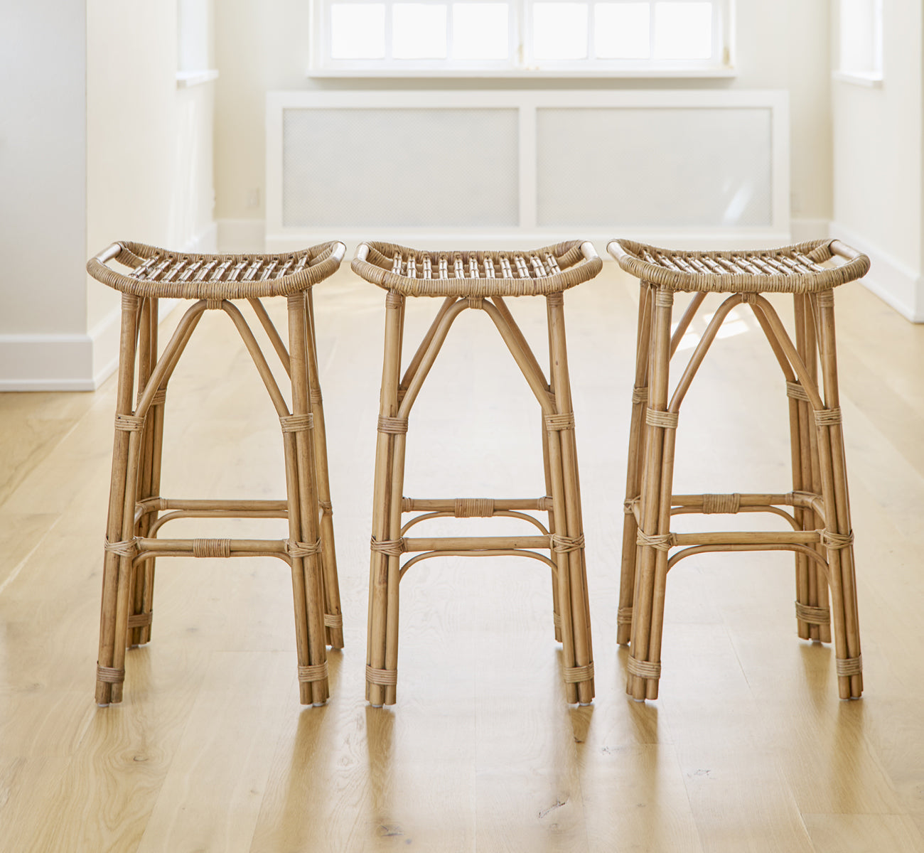 This is a lifestyle image of Salsa Bar Stool in antique rattan. A pair placed against a white wall on a herringbone parquet floor.
