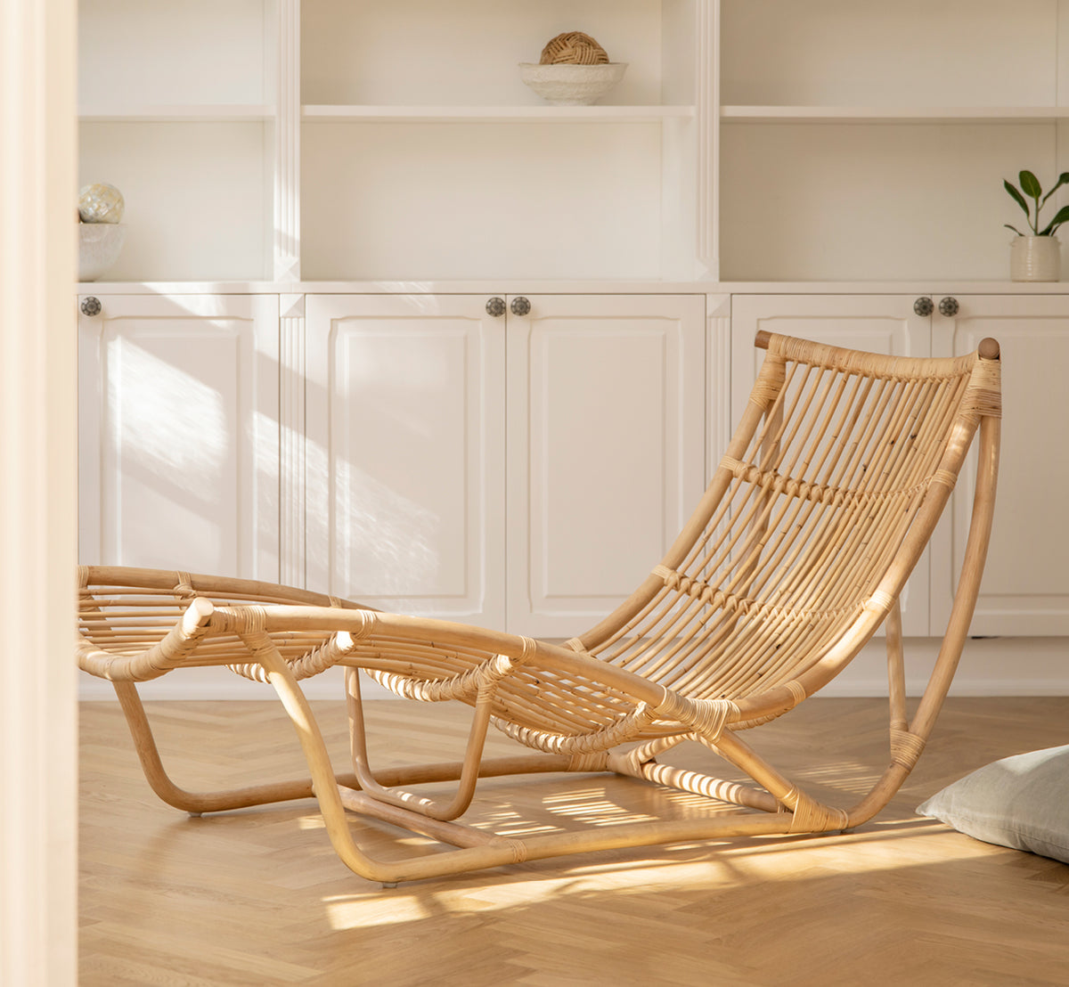 This is a lifestyle image of Michelangelo Daybed in natural rattan. Placed in a bright white room with concrete floor and pink lamp.