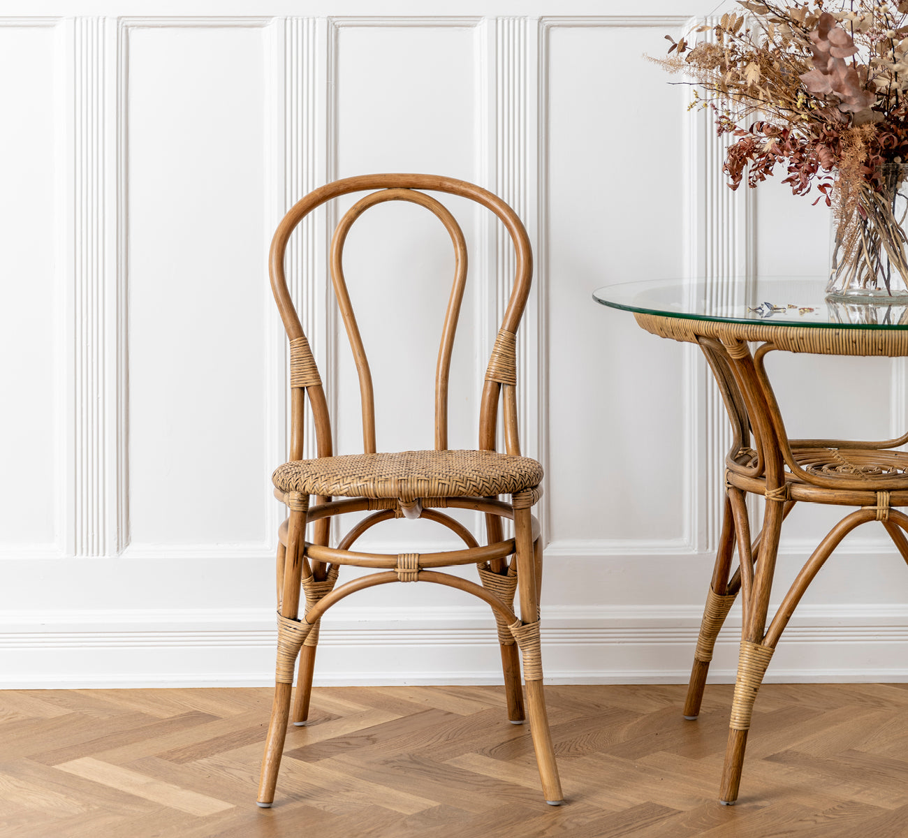 This is a lifestyle image of Lulu Dining Chair in antique rattan. Placed beside a glass rattan table with dried flowers in a paneled interior.