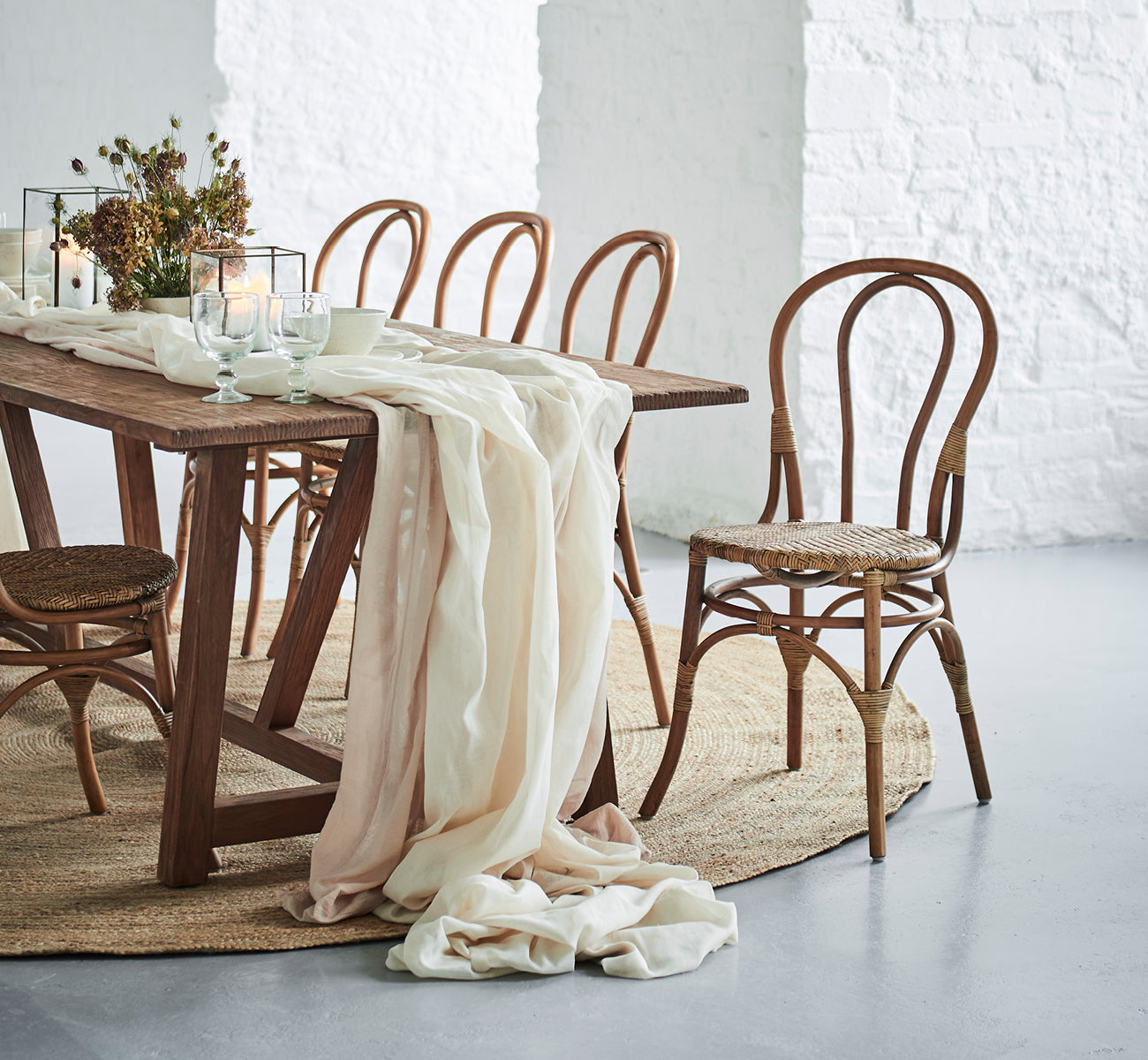 This is a lifestyle image of Lulu Dining Chair in antique rattan. Set at a rustic table with linen draping and candles against a white brick wall.