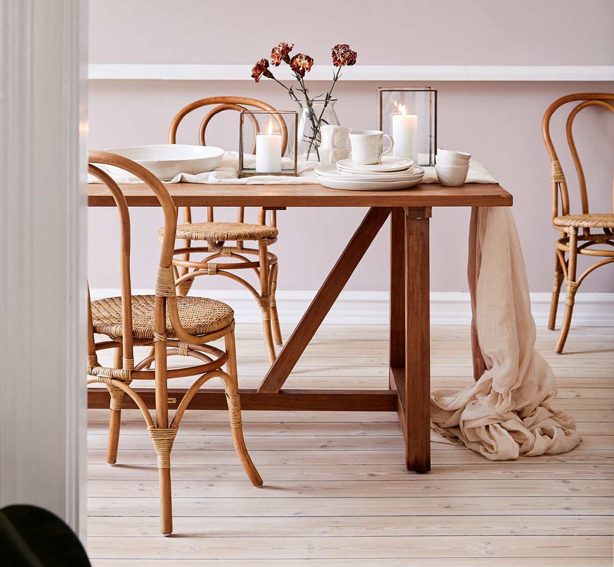 This is a lifestyle image of Lulu Dining Chair in antique rattan. Styled at a teak dining table with candles and linen in a warm-toned interior.