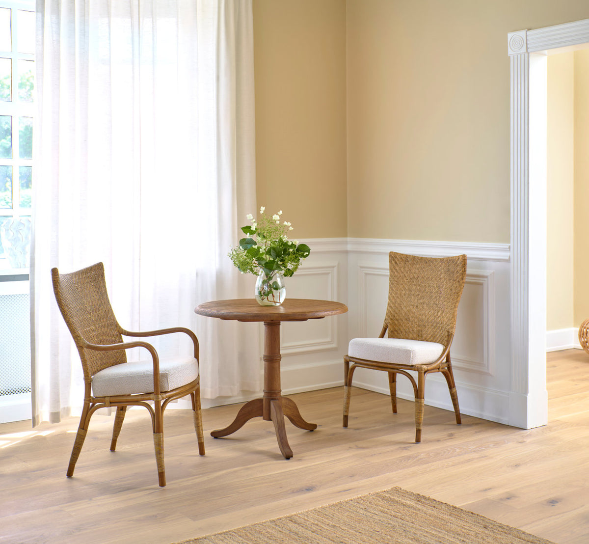 This is a lifestyle image of Melody Dining Armchair in antique rattan. Pair at a teak café table by white panels.