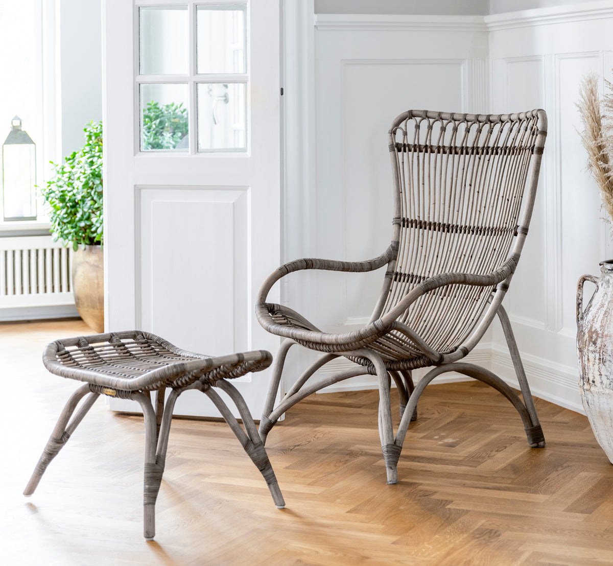 This is a lifestyle image of Monet Footstool in taupe rattan. Paired with lounge chair in a dark barn setting with candle and glass.