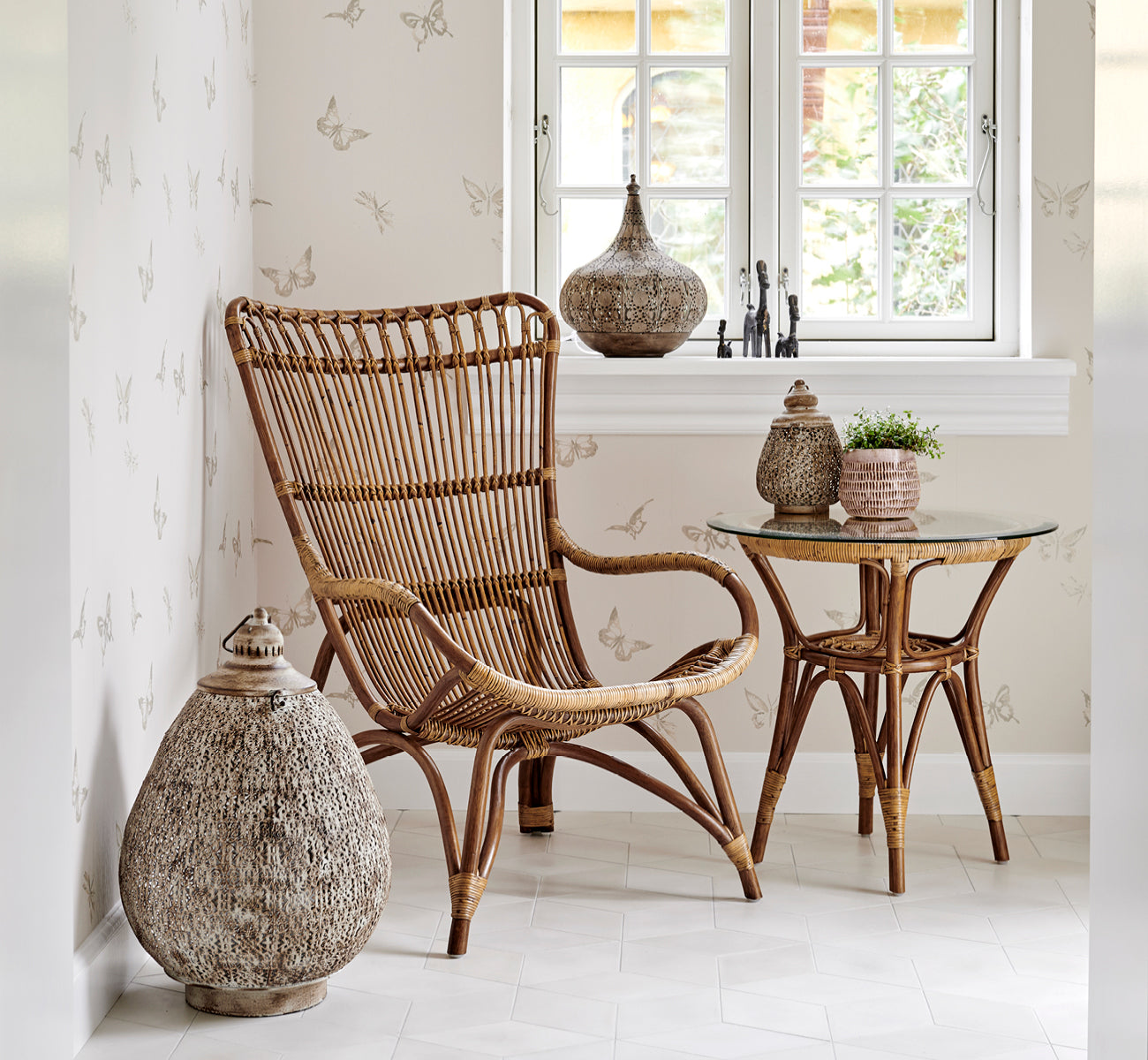 This is a lifestyle image of Tony Side Table in antique rattan. Set in a butterfly-print wallpapered corner with a lantern and decor.