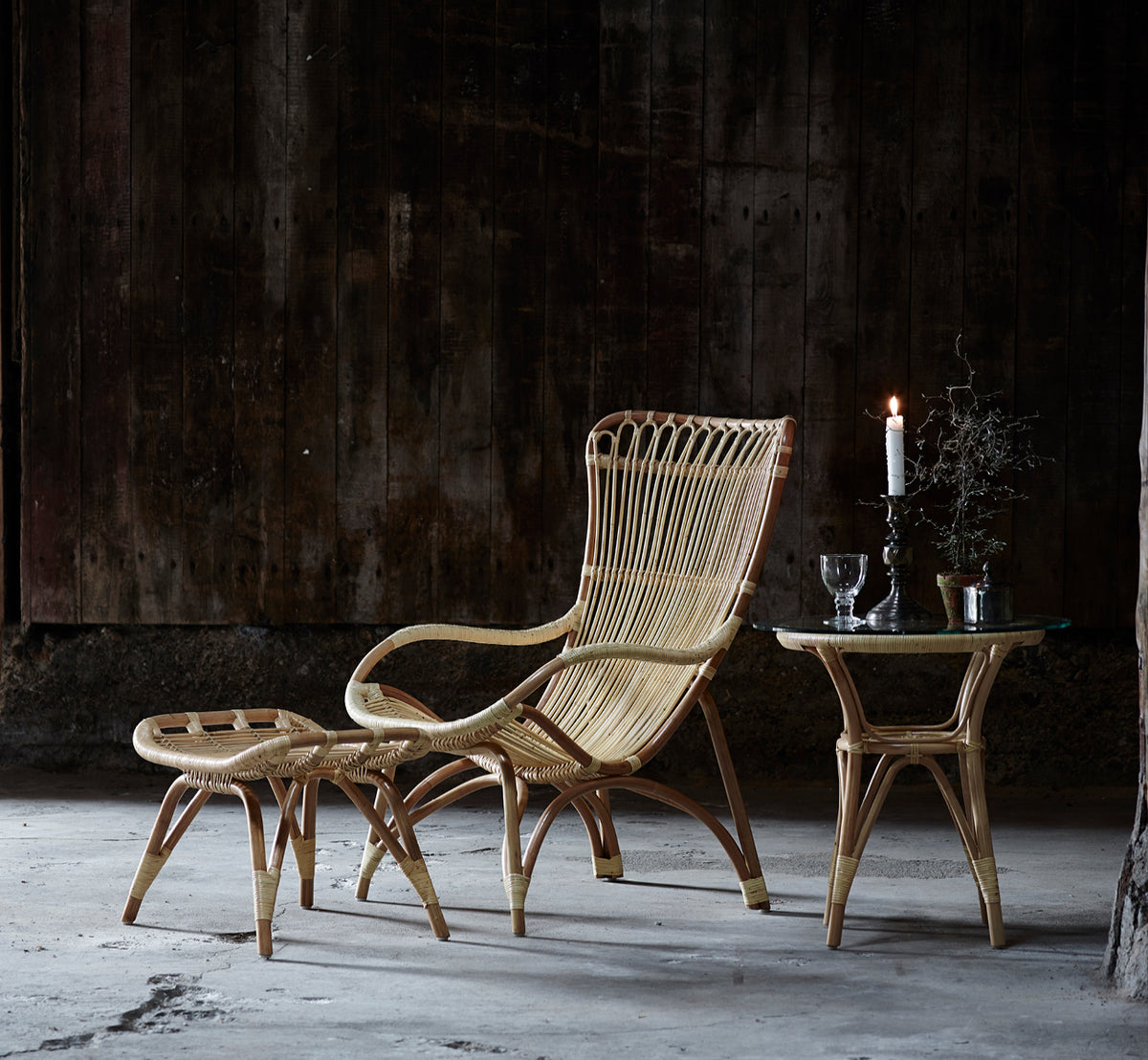This is a lifestyle image of Tony Side Table in natural rattan. Paired with a high-back lounge chair against a dark wood wall.