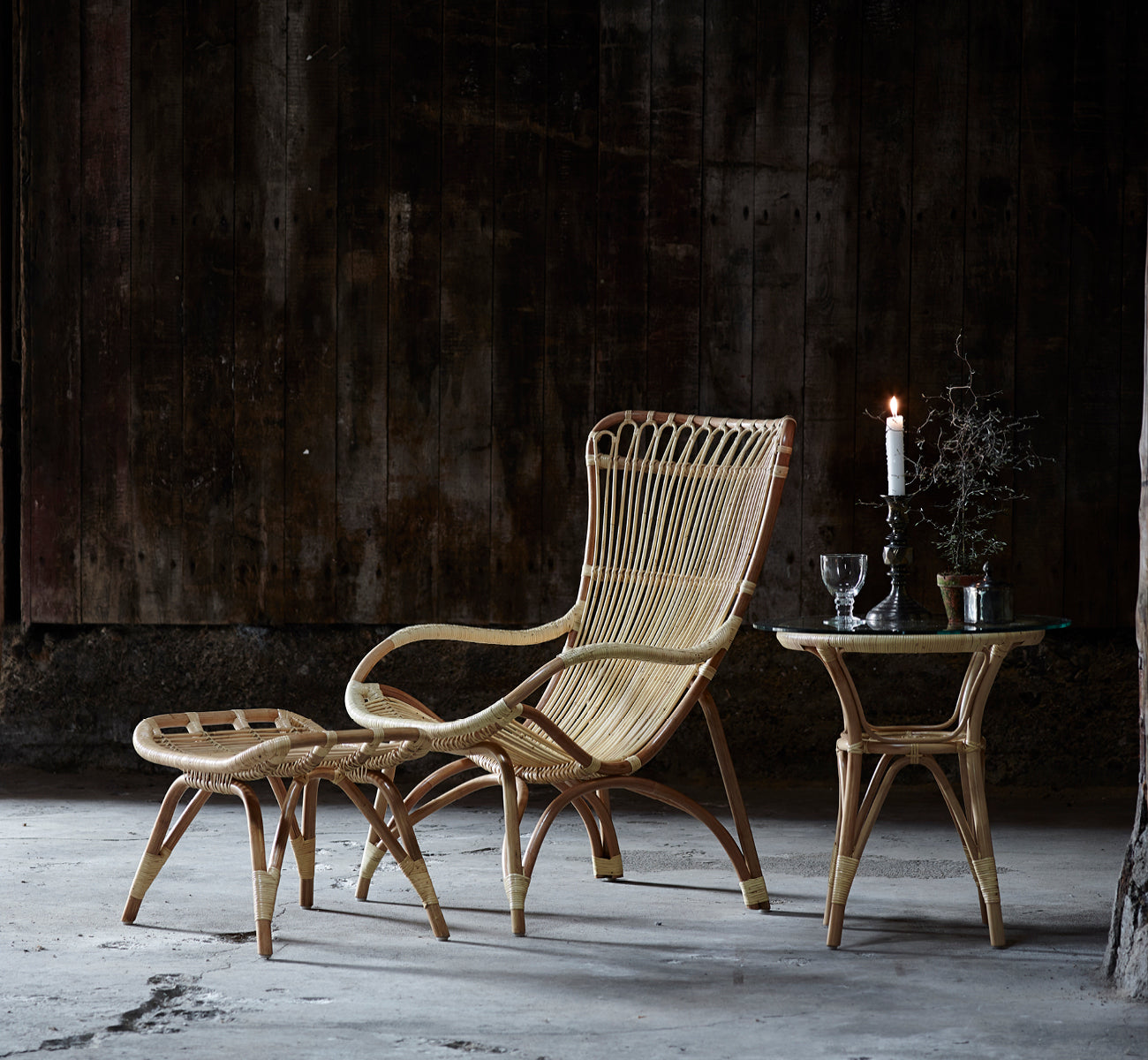 This is a lifestyle image of Tony Side Table in natural rattan. Paired with a high-back lounge chair against a dark wood wall.