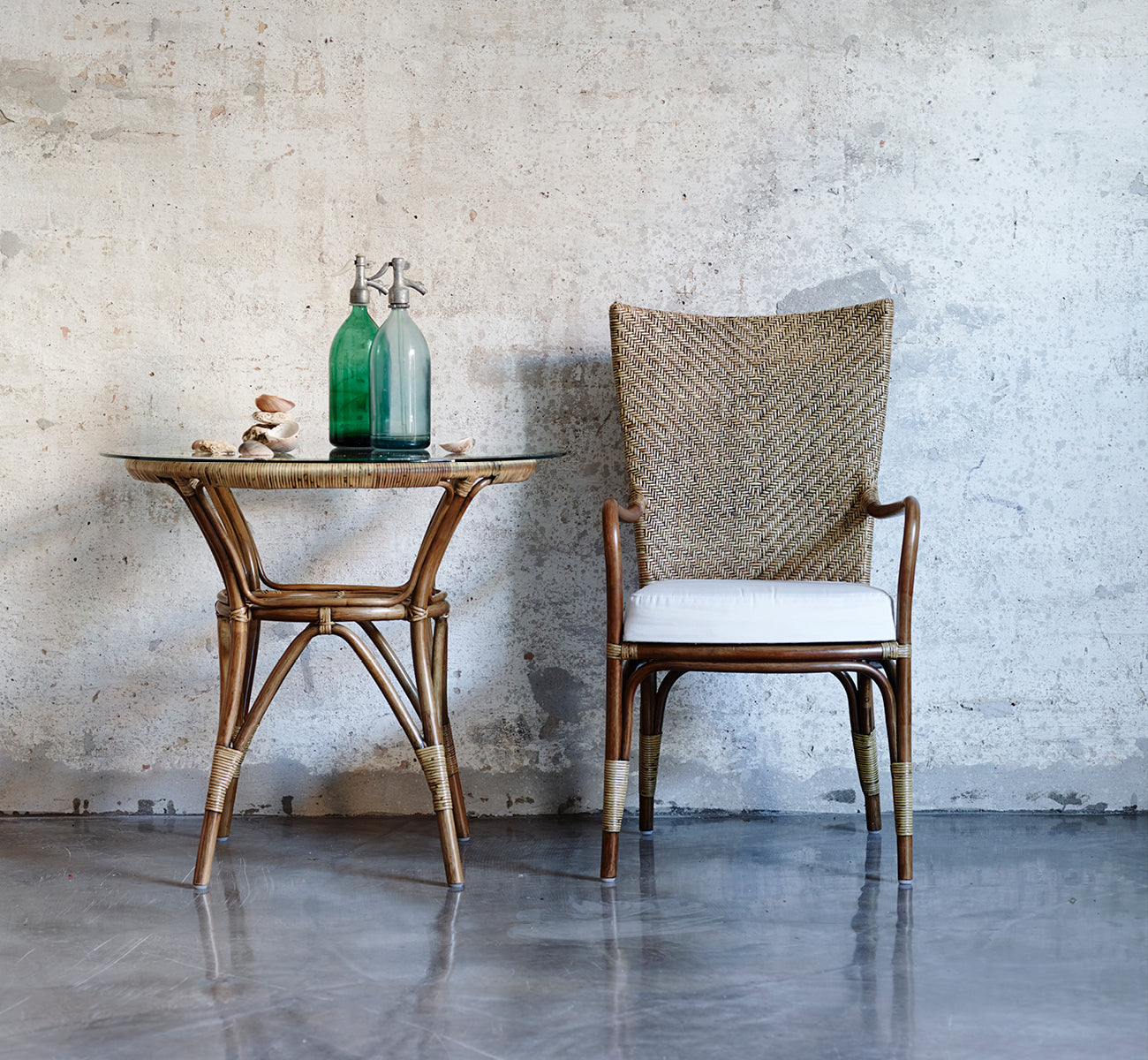 This is a lifestyle image of Tony Café Table in antique rattan. Paired with a high-back chair against a raw concrete wall.