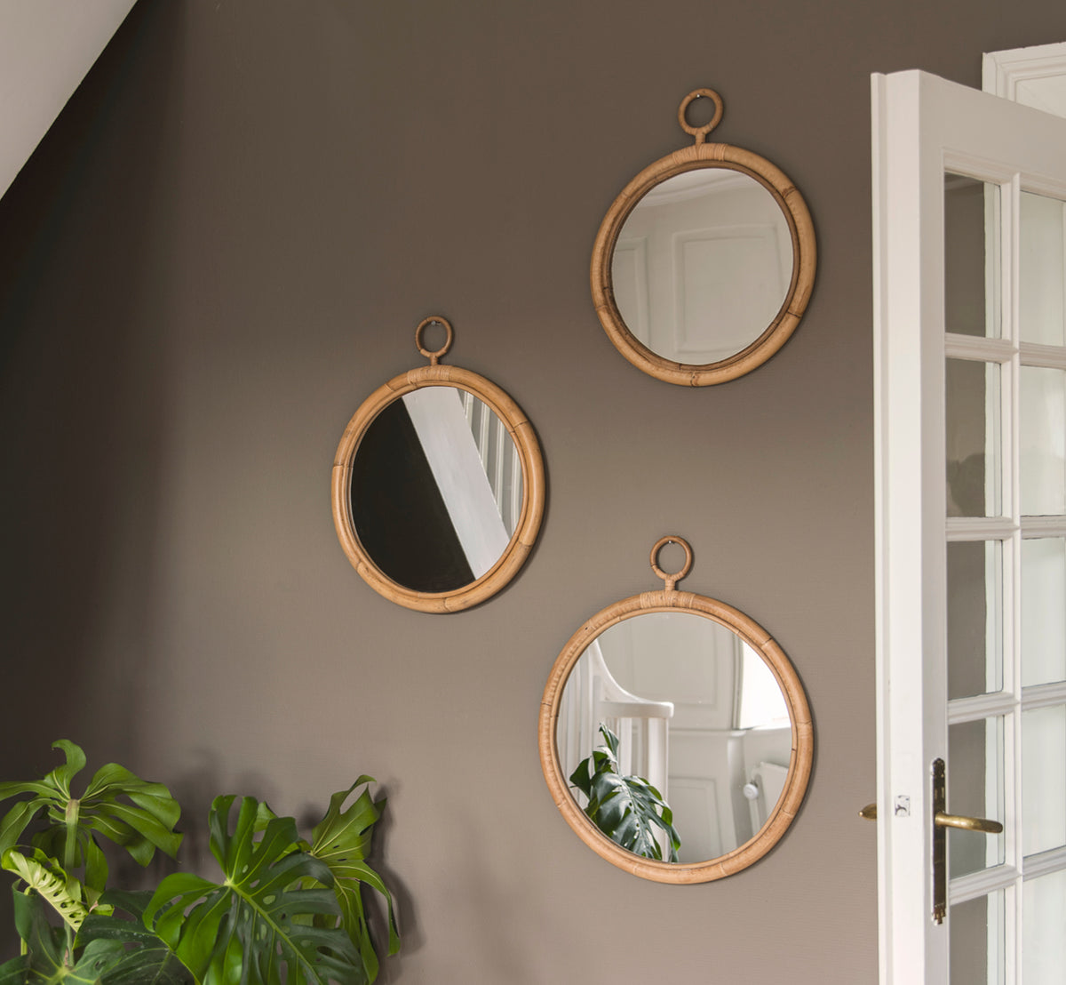 This is a lifestyle image of Ella Mirror O45cm in natural skin on rattan, on a taupe wall in a hallway with a monstera plant and white door.
