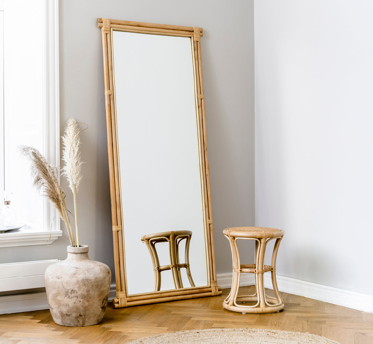 This is a lifestyle image of Rasmus Mirror 172 cm in natural rattan. Leaning against a grey wall beside a stool and dried pampas grass.