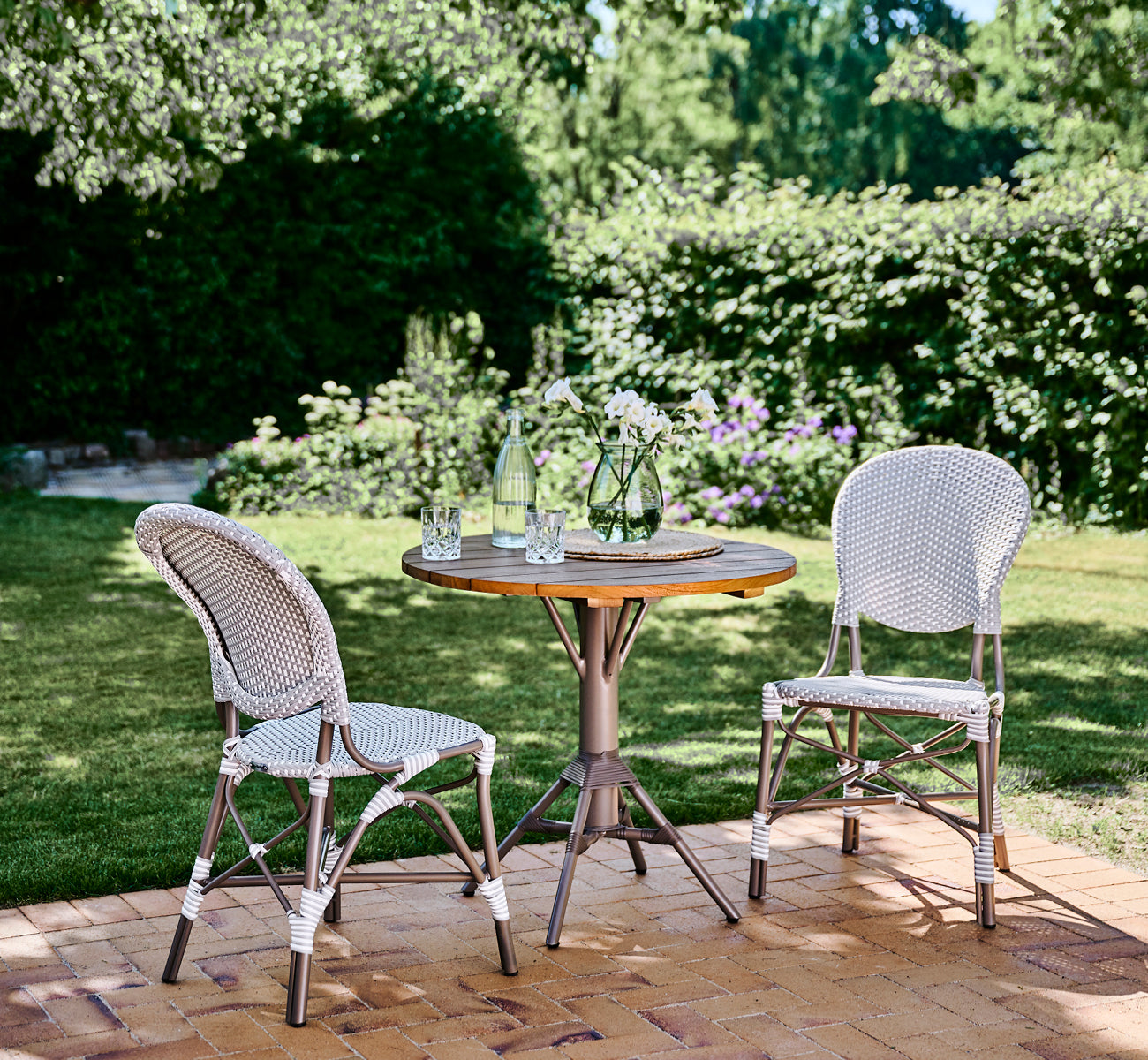 This is a lifestyle image of Isabell in taupe aluminium frame Grey w white dot ArtFibre, in a lush garden patio with stone tile and a greenery-clad.