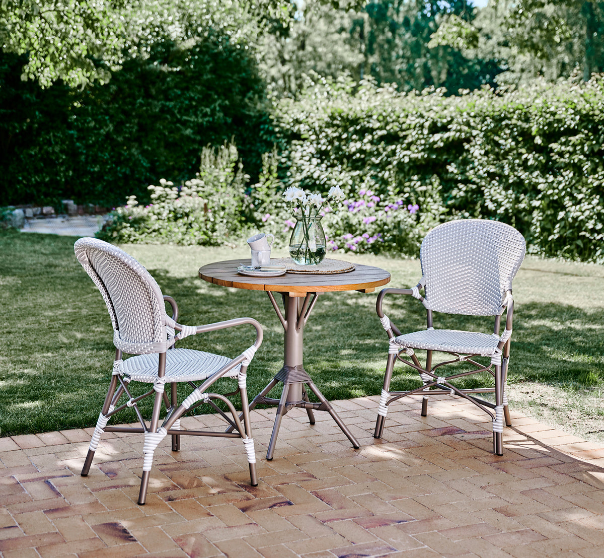 This is a lifestyle image of Isabell in taupe aluminium frame Grey w white dot ArtFibre, in a lush garden patio with stone tile and a greenery-clad.