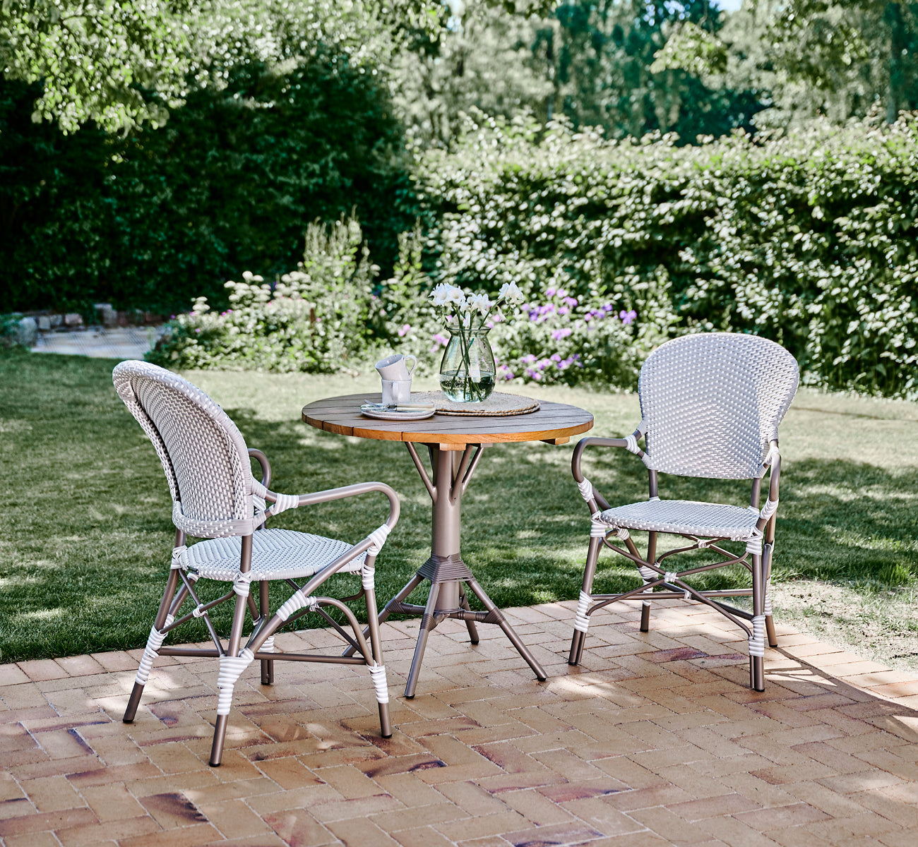 This is a lifestyle image of Isabell in taupe aluminium frame Grey w white dot ArtFibre, in a lush garden patio with stone tile and a greenery-clad.