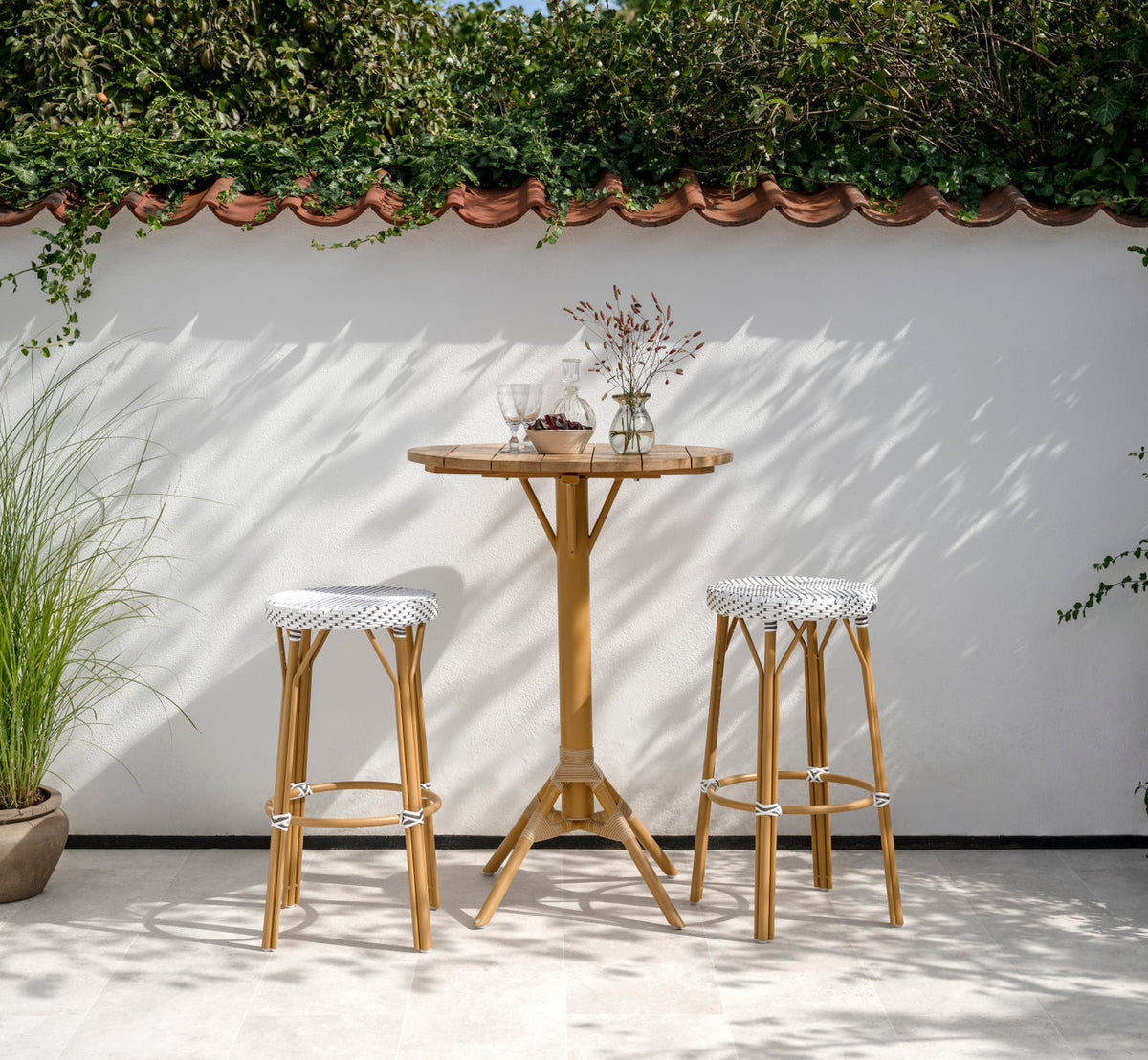 This is a lifestyle image of Simone Exterior Bar Stool in almond aluminium frame white w cappuccino dot artfibre. At a teak bar table on a terrace.