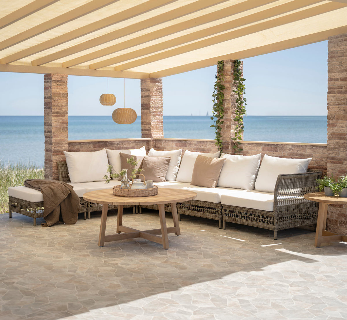 This is a lifestyle image of Mona Exterior Modular Sofa-Center Module in antique grey artfibre aluminium. Under a pergola by the sea.