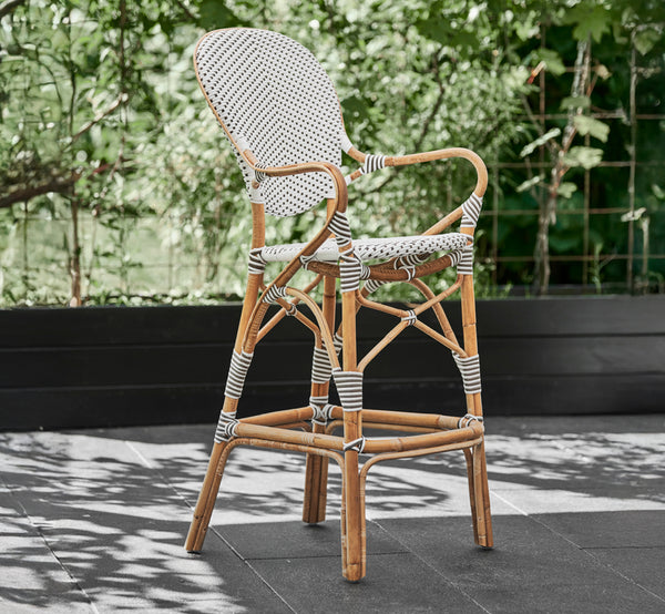 This is a lifestyle image of Isabell Bar Stool in white w cappuccino dot ArtFibre Skin on Rattan frame, outdoors against a lush green garden hedge on.