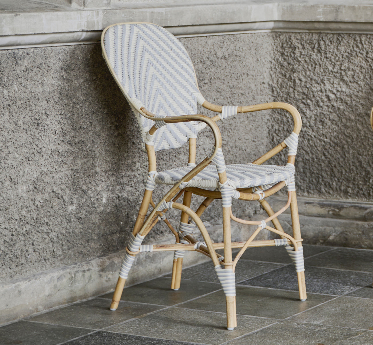 This is a lifestyle image of Isabell Dining Armchair in chevron Grey Ivory ArtFibre Skin on Rattan frame, in a bright dining area with large windows.