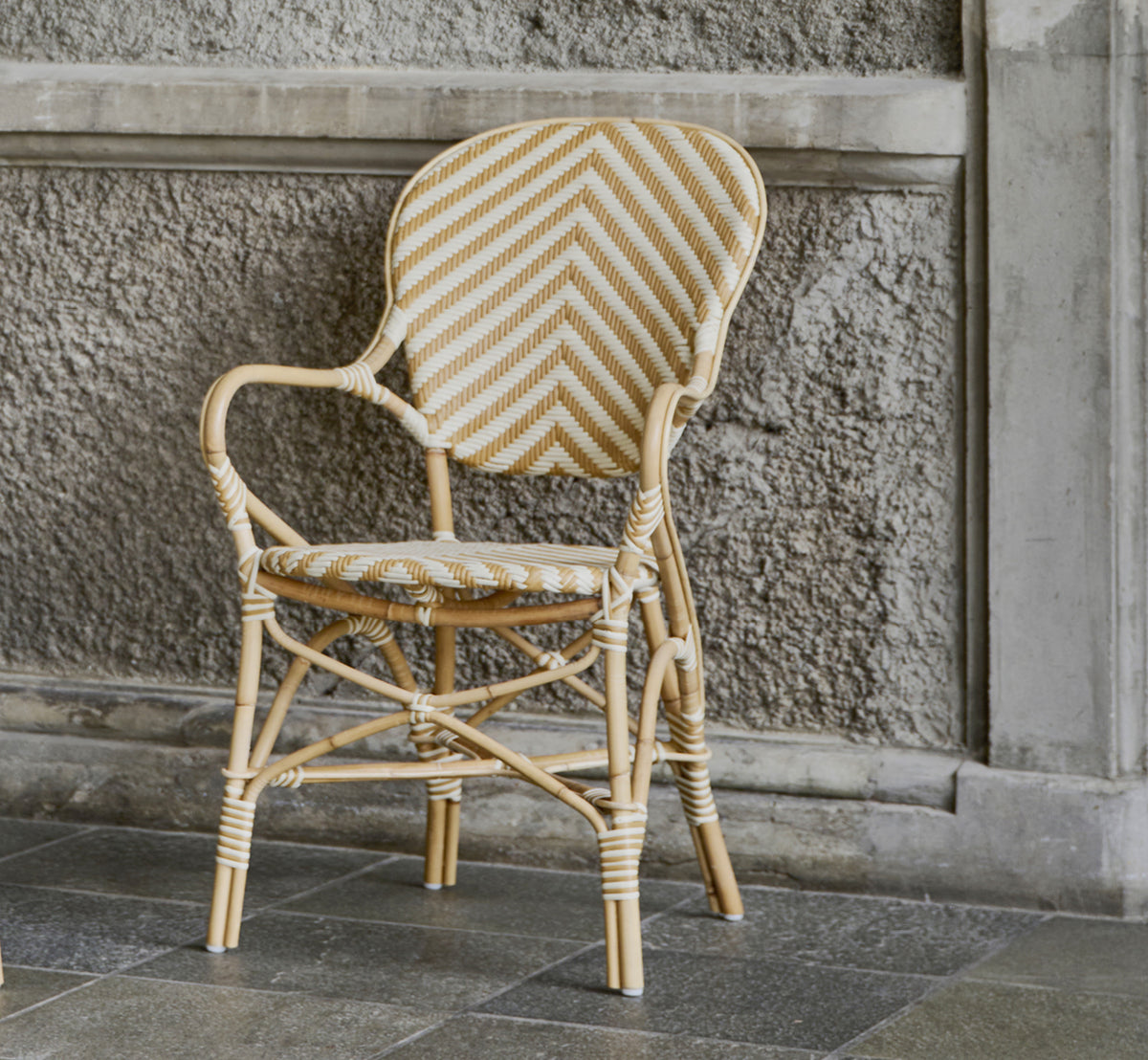 This is a lifestyle image of Isabell Dining Armchair in chevron Honey Ivory ArtFibre Skin on Rattan frame, in a bright dining area with large windows.
