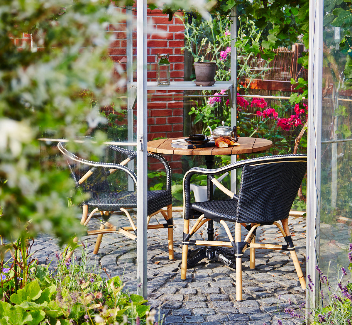 This is a lifestyle image of Madeleine Dining Chair in black ArtFibre skin on rattan frame. Garden greenhouse with cobblestone floor and plants.