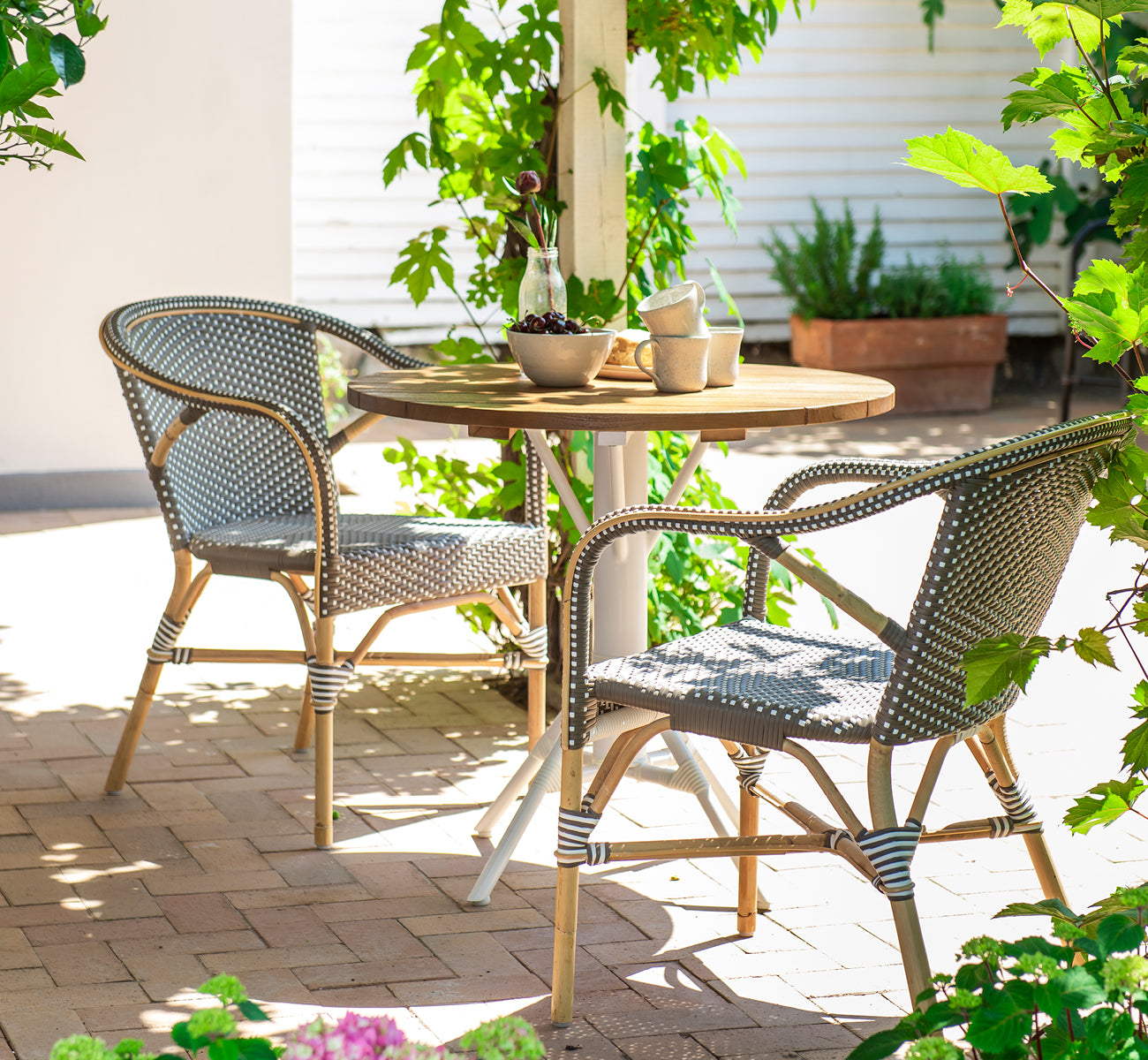 This is a lifestyle image of Madeleine Dining Chair in cappuccino w white dot rattan frame. Shaded garden terrace with round teak table.