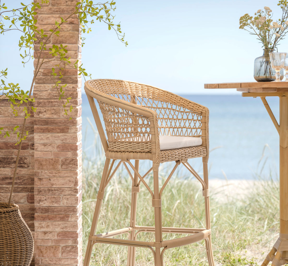 This is a lifestyle image of Emma Exterior Barstool in natural artfibre aluminium. By a brick pillar with dune grass and sea view.
