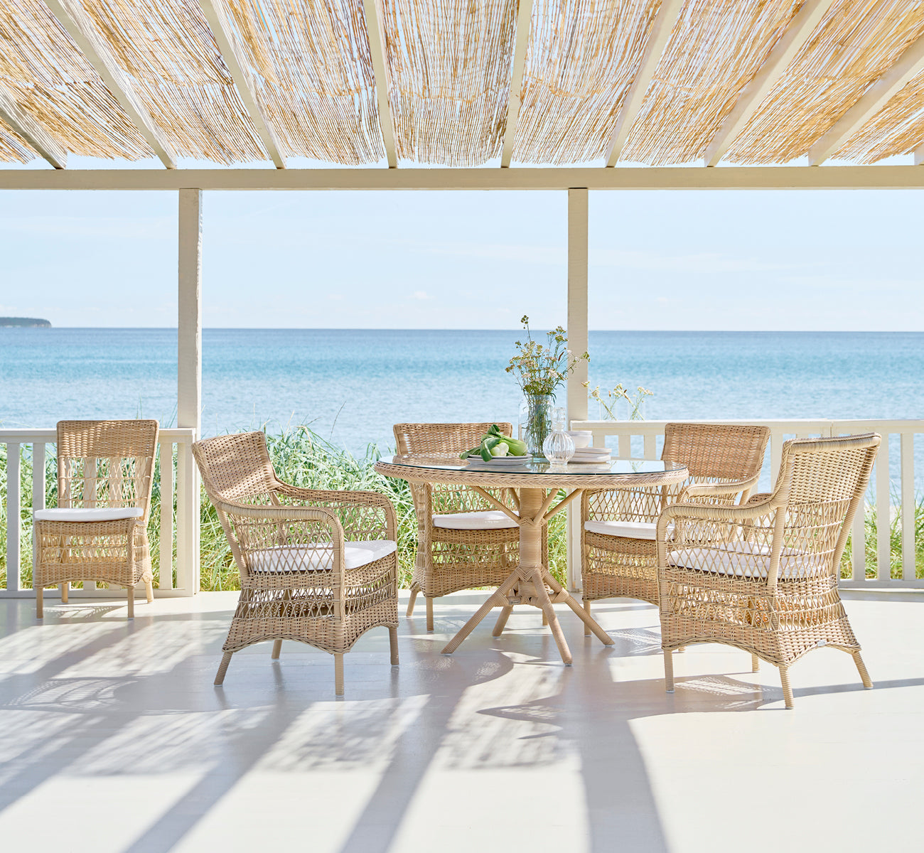 This is a lifestyle image of Marie Exterior Armchair in natural artfibre aluminium. On a covered veranda with seaside view.