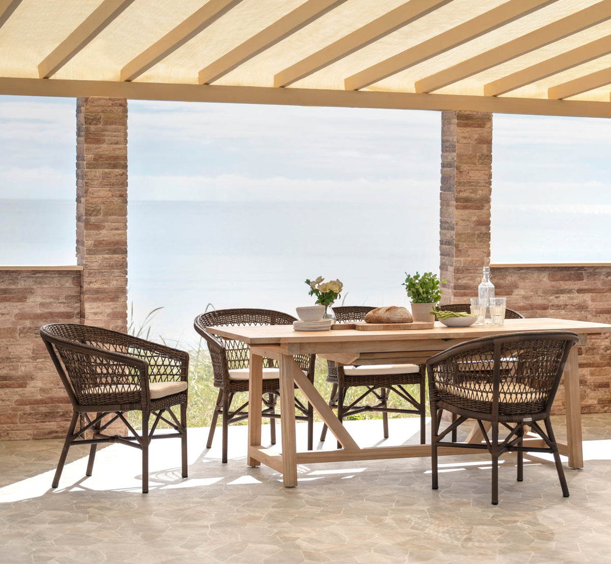 This is a lifestyle image of Emma Exterior Chair in brown artfibre aluminum. Set for lunch on a terrace by the sea.