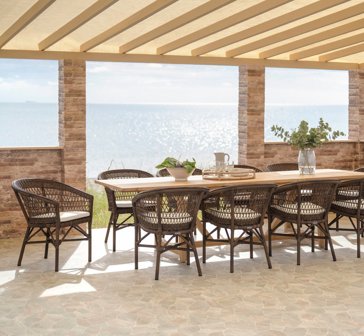 This is a lifestyle image of Emma Exterior Chair in brown artfibre aluminum. Long dining setting on a terrace by the sea.