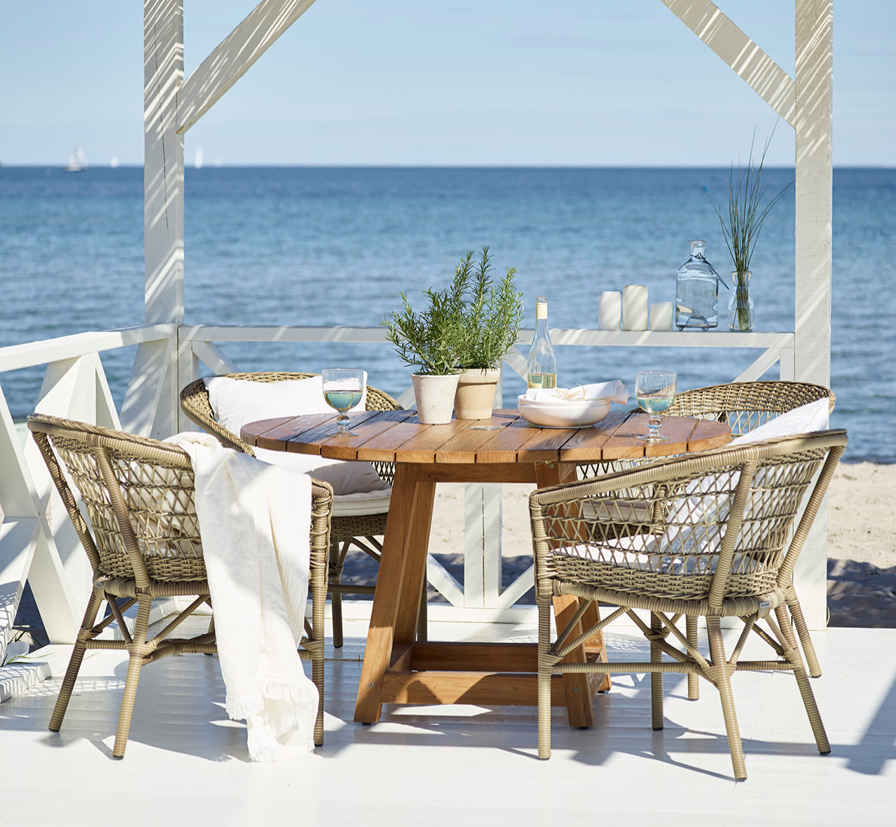 This is a lifestyle image of Emma Exterior Dining Chair in antique Grey ArtFibre Aluminium, on a bright coastal terrace with a reed roof shade and.