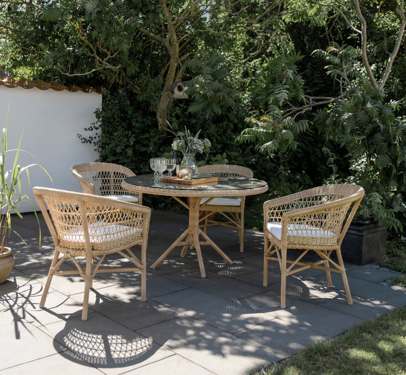 This is a lifestyle image of Emma Exterior Dining Chair in natural ArtFibre Aluminium, on a bright coastal terrace with a reed roof shade and sea.