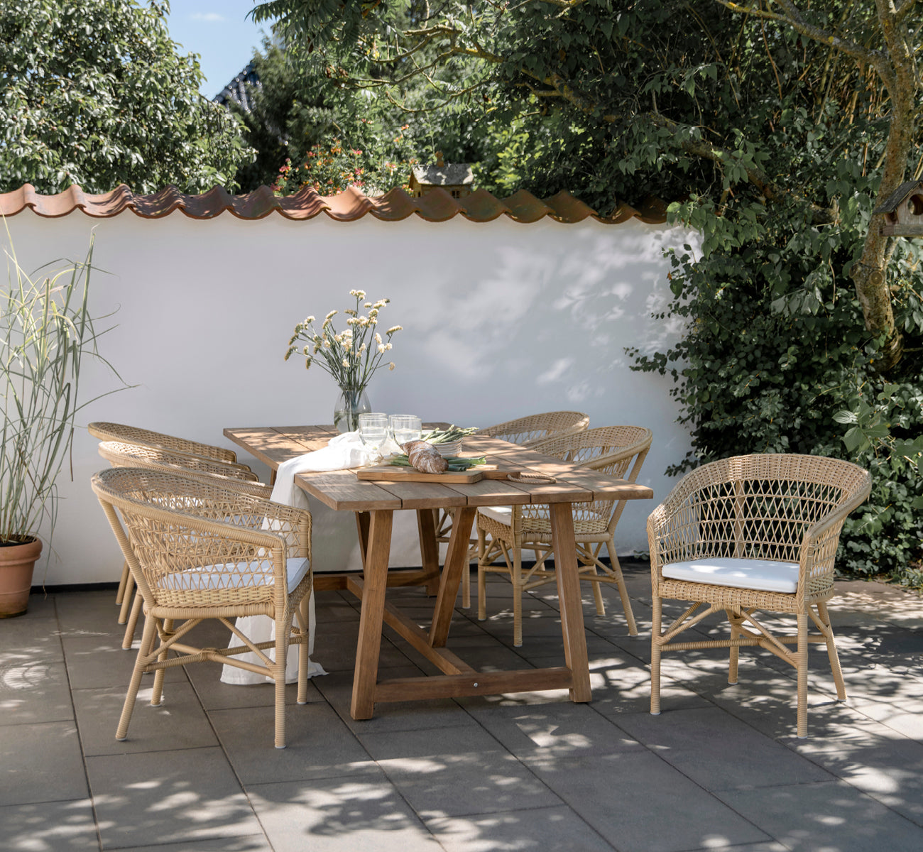 This is a lifestyle image of Emma Exterior Dining Chair in natural ArtFibre Aluminium, on a bright coastal terrace with a reed roof shade and sea.