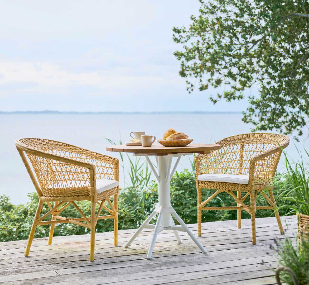 This is a lifestyle image of Emma Exterior Dining Chair in natural ArtFibre Aluminium, on a bright coastal terrace with a reed roof shade and sea.