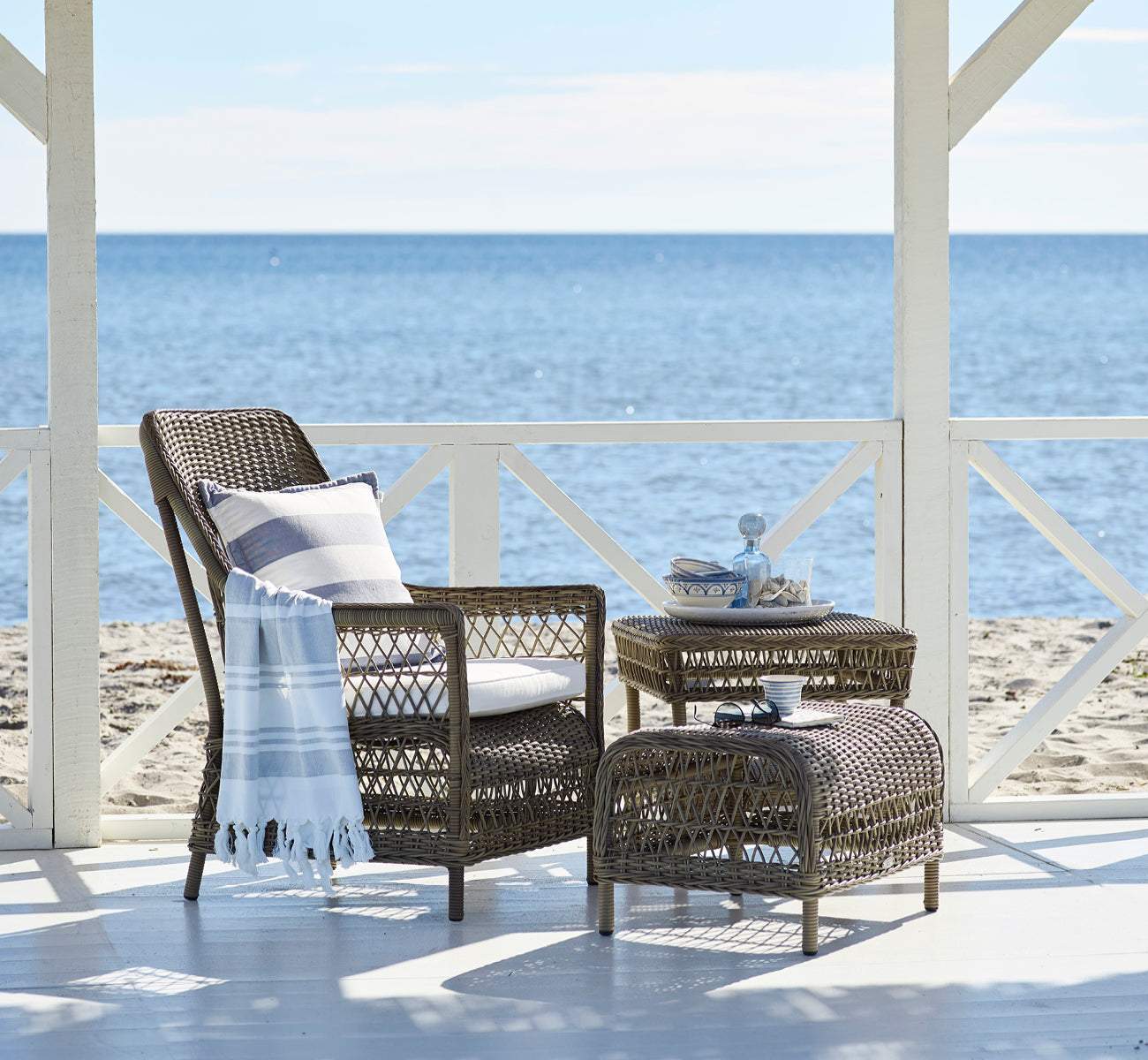 This is a lifestyle image of Dawn Lounge Chair in antique Grey ArtFibre Aluminium, on a coastal terrace with sea views, beach grasses and white.