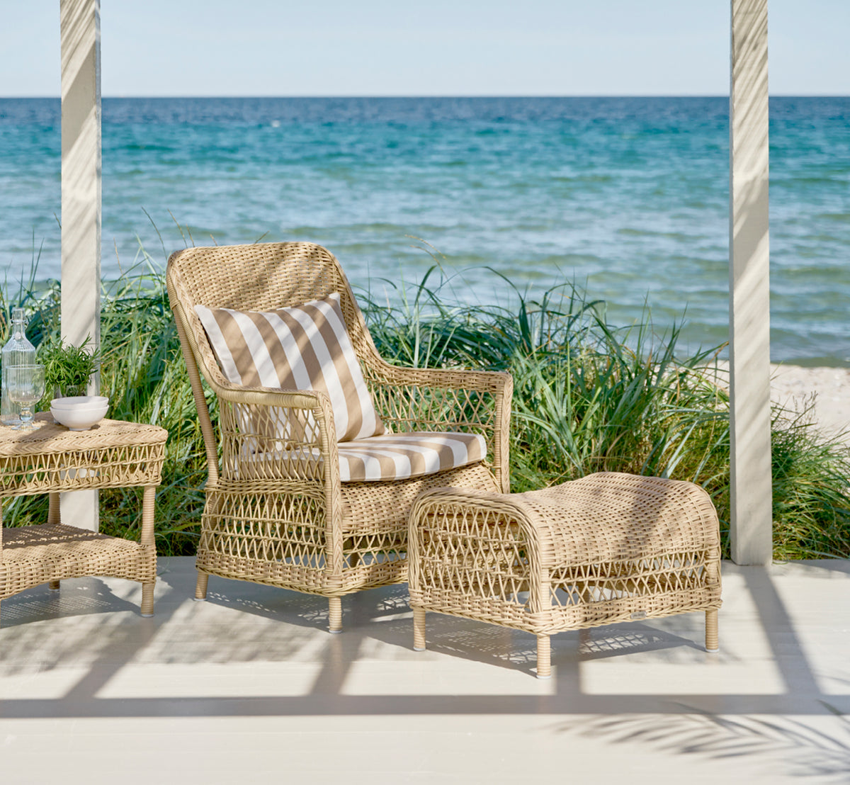 This is a lifestyle image of Dawn Lounge Chair in natural ArtFibre Aluminium, on a coastal terrace with sea views, beach grasses and white columns.