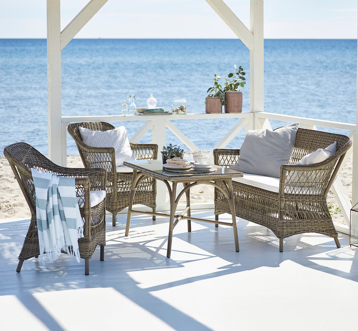 This is a lifestyle image of Charlot 2 Seater Sofa in antique Grey ArtFibre Aluminium, on a coastal terrace with reed roof, sea views and striped.