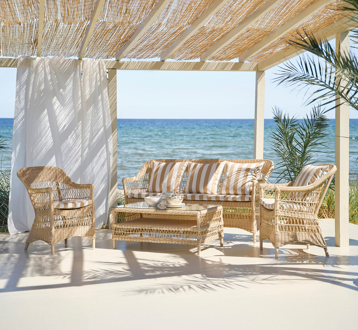 This is a lifestyle image of Charlot 3 Seater Sofa in natural ArtFibre Aluminium, on a coastal terrace with reed roof shade and sea views.