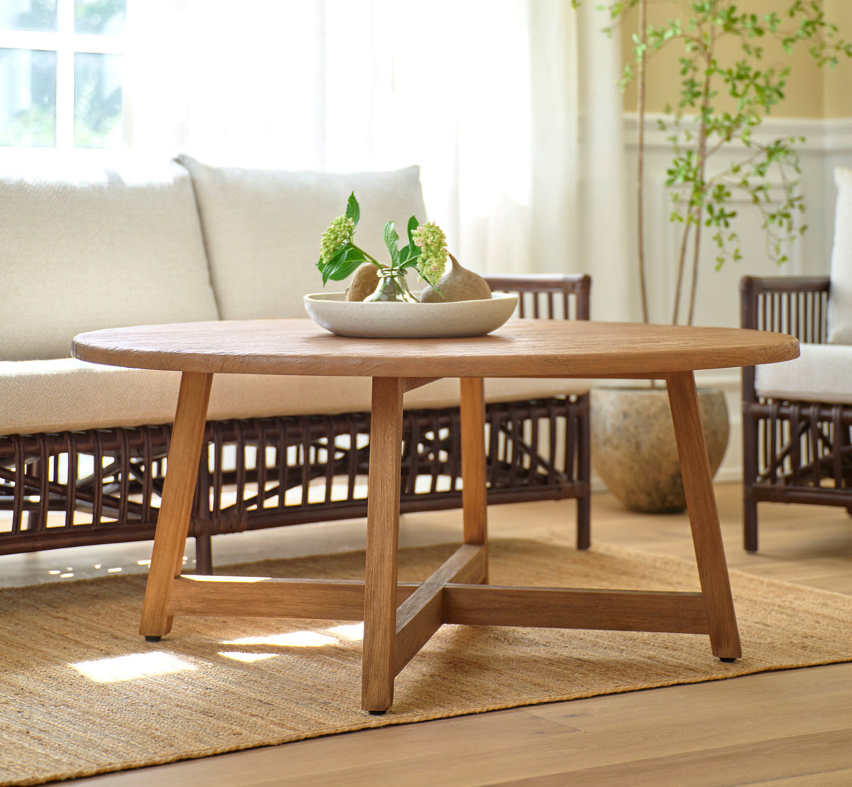 This is a lifestyle image of Aksel Coffee Table Ø120 cm in teak indoor. Before a rattan sofa in a sunny living room.